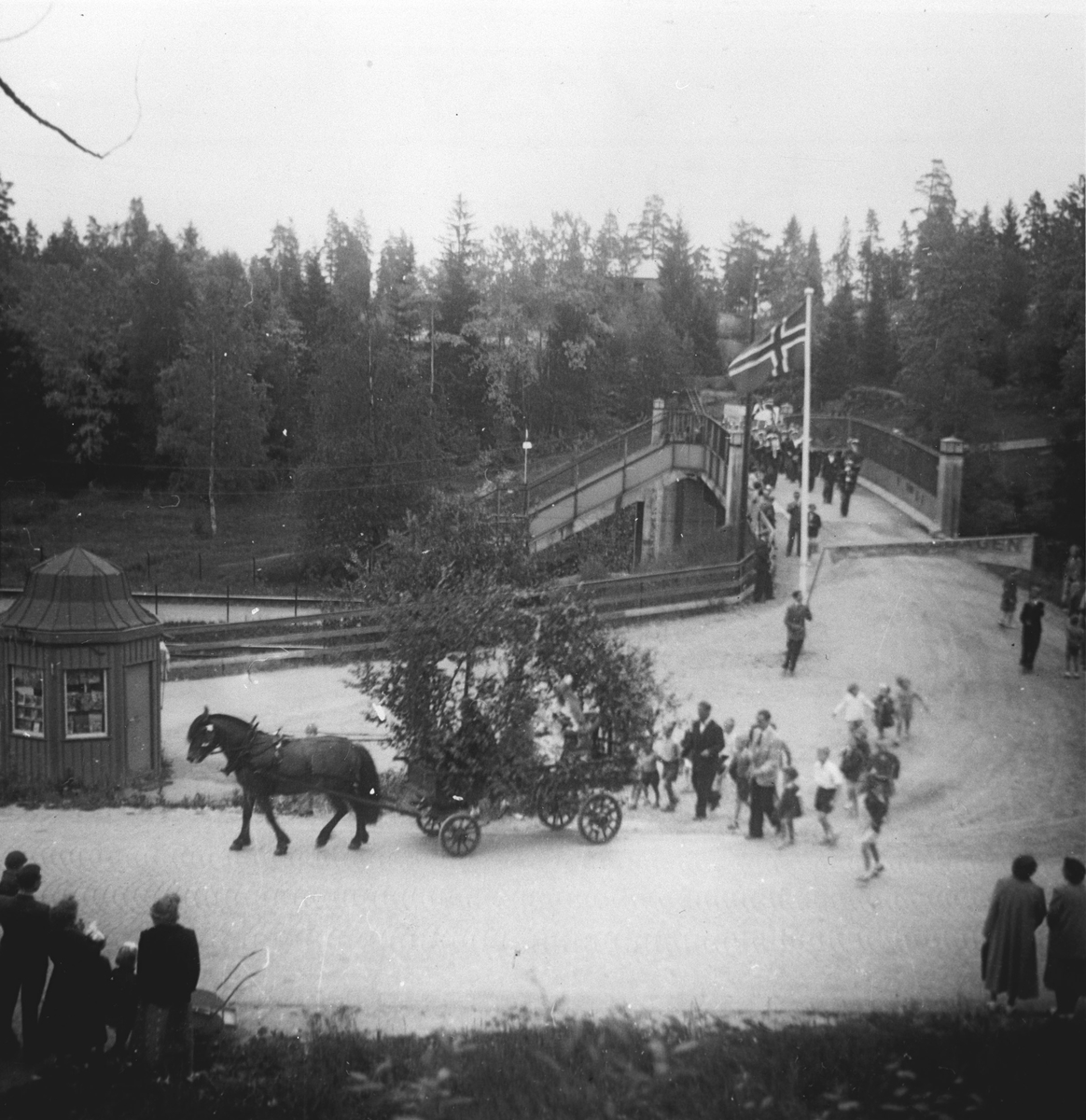 Musikkorps passerer over Myrvollbrua. Hest med løvpyntet kjerre passerer flaggstanga og kiosken.