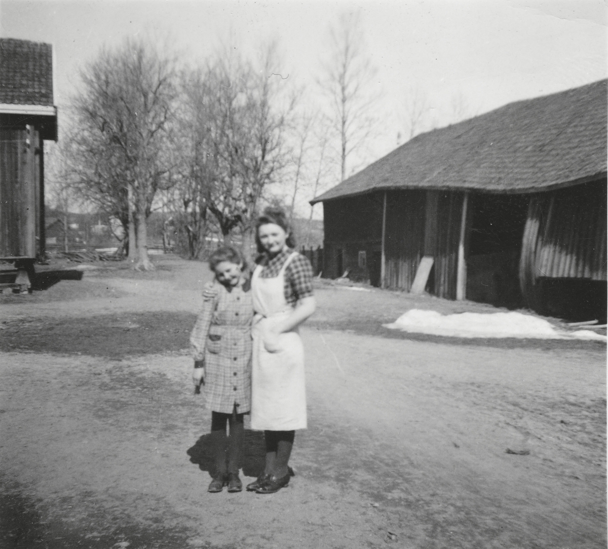 Ingrid Frogner og Liv Ragnhild Frogner Knutsen står ute på gårdstunet, foran den gamle høylåven og stabburet på Frogner gård.