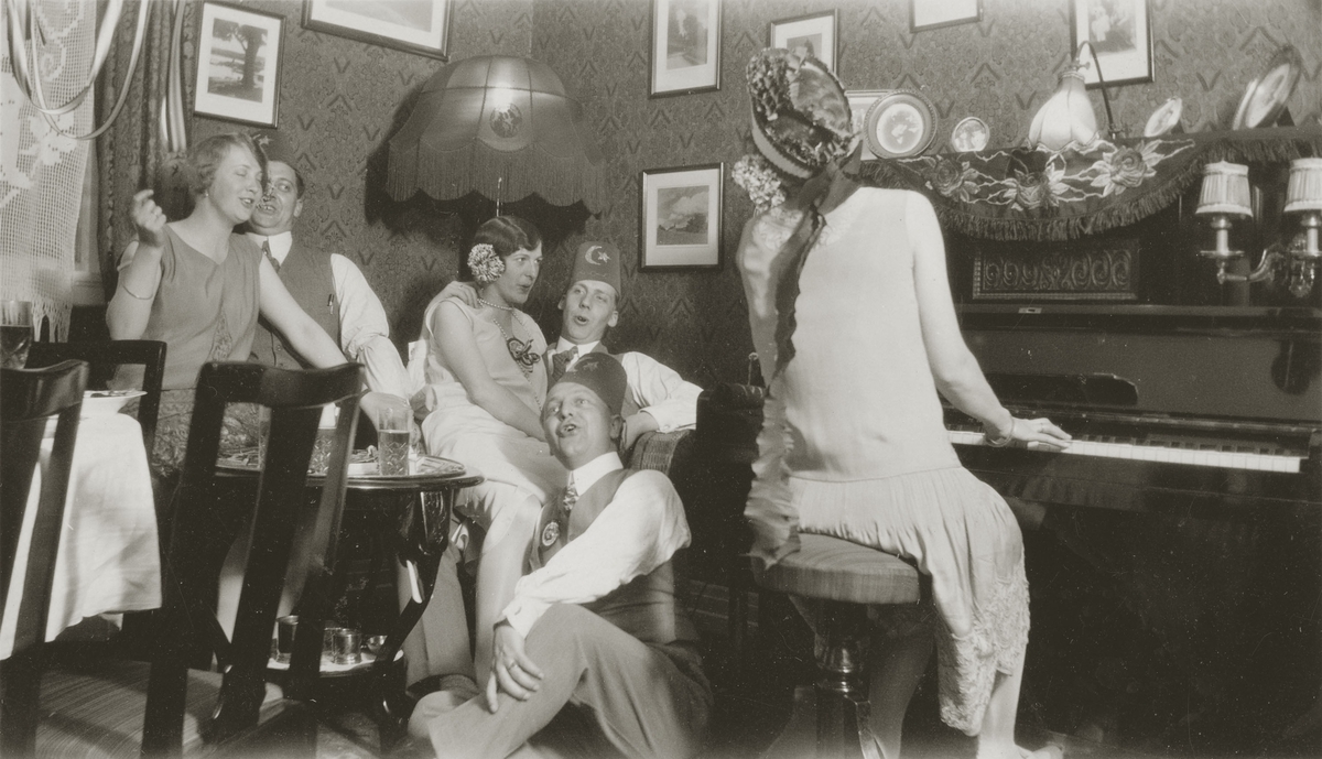 En gruppe kvinner og menn i festlig lag. En kvinne spiller på piano av typen  H. Wolfframm Dresden. Party hatter.