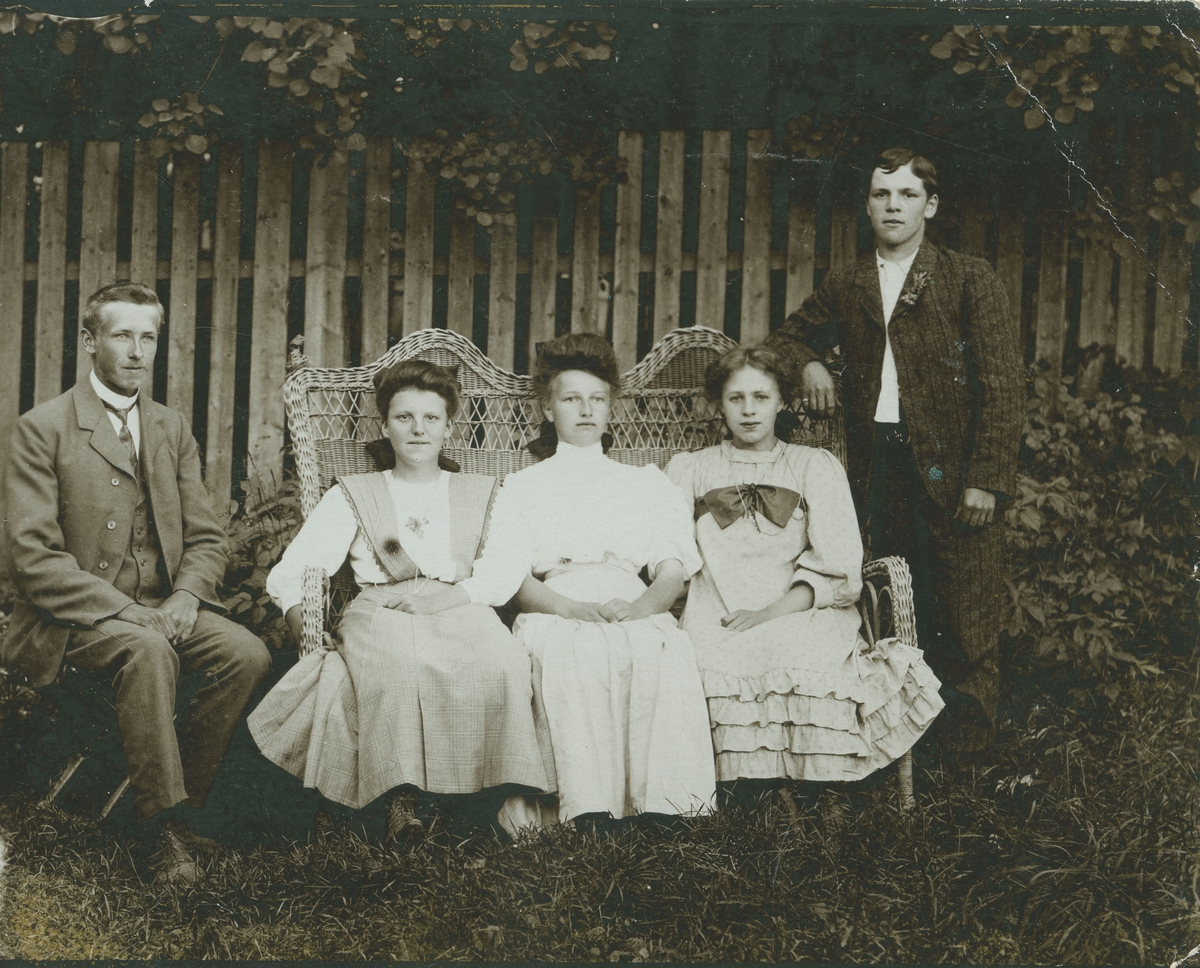 Gruppebilde av to unge menn og tre unge damer som sitter i kurvflettet hagebenk.
