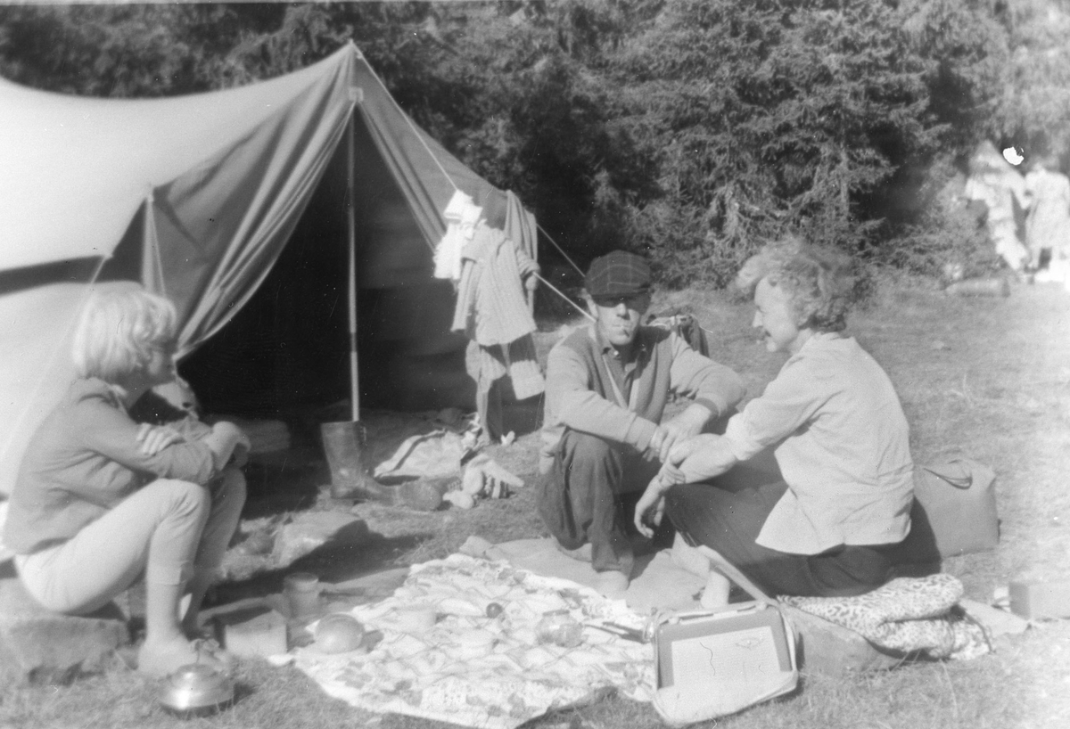 Camping, telttur. Måltid inntas foran teltet med reiseradio som underholdning.