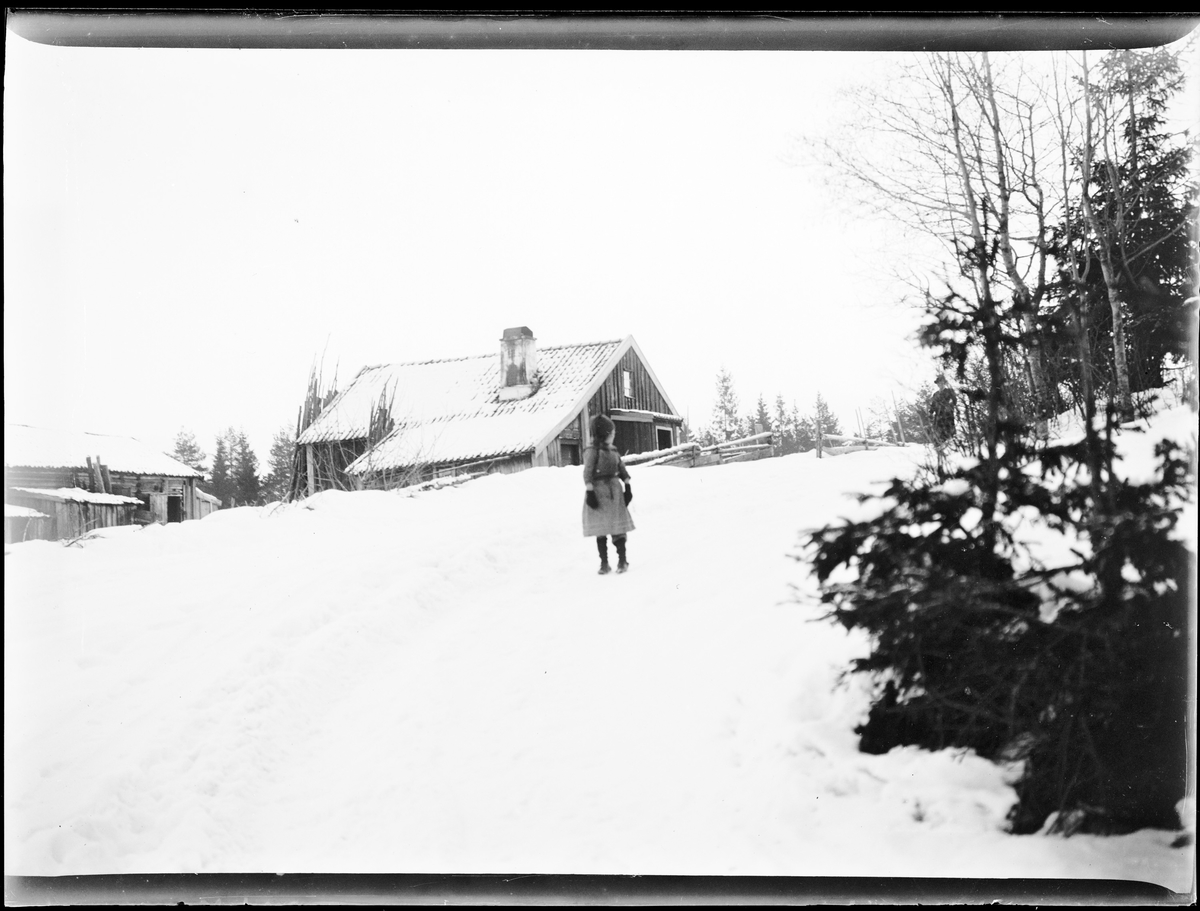 Vinterlandskap. En jente midt i bildet. Hus/hytte i bakgrunnen.