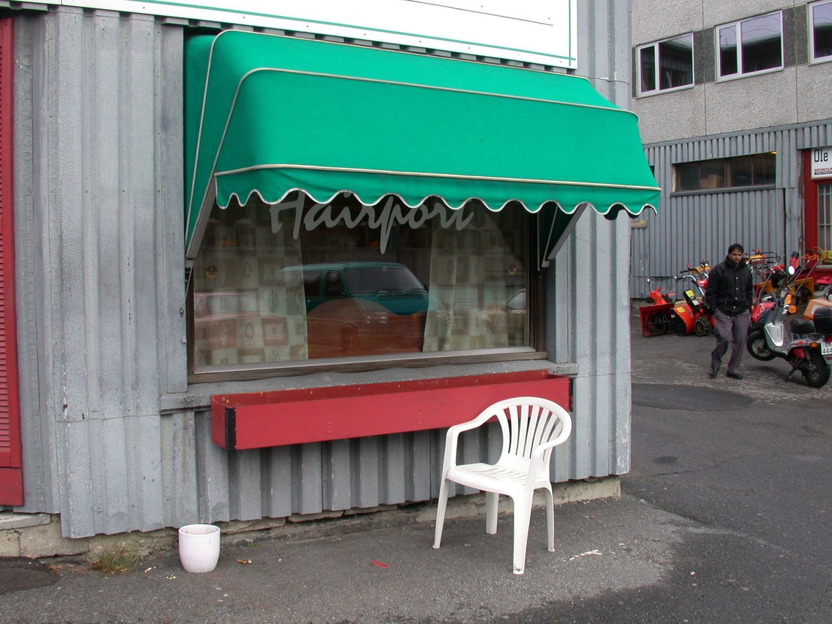 Gamle Ra-Glabygget og en plaststol og potteskjuler for utendørs røyking. Utenfor frisøren.
Fotovinkel: N