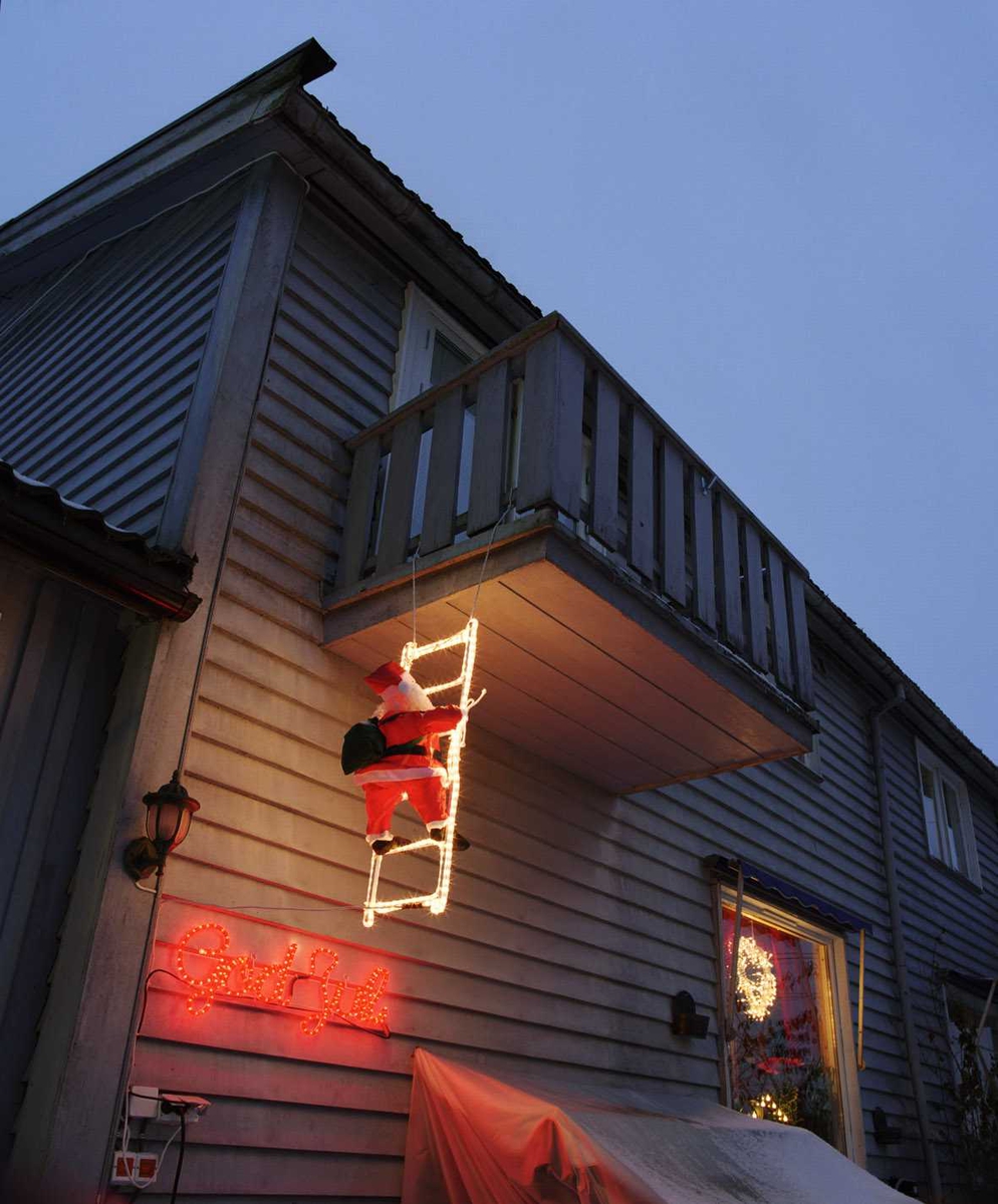 Julebelysning.  

Klatrende nisse og rød lysende skrift med god jul på husvegg på rekkehus. Lysende krans i vindu