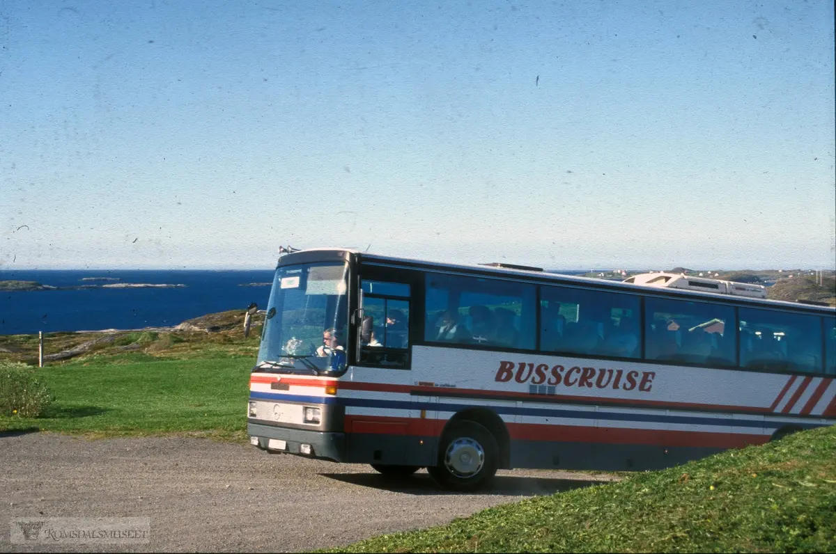 Turistbuss ankommer Ergan i Bud...Denne bussen er lakkert i fargene til Nordmøre Trafikkselskap A/S (NTS) som var resultat av en fusjon fra 1. januar 1987. Bussen er også merket "Busscruise". Dette var navnet på et samarbeid mellom Kristiansund Frei Billag (KFB) og Molde Bilruter om feriereiser med buss. Året etter ble også Ålesund Bilruter med i "Busscruise". KFB ble med i NTS fra 1987 og bildet er derfor tidligst fra dette året.