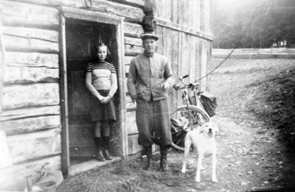 Kolbjørn Mathiasen Grøtlien (1898-1964) med hund utenfor huset i Granerud, Nes, Hedmark. Kari f.1937 i strikket genser og skjørt står i døra.