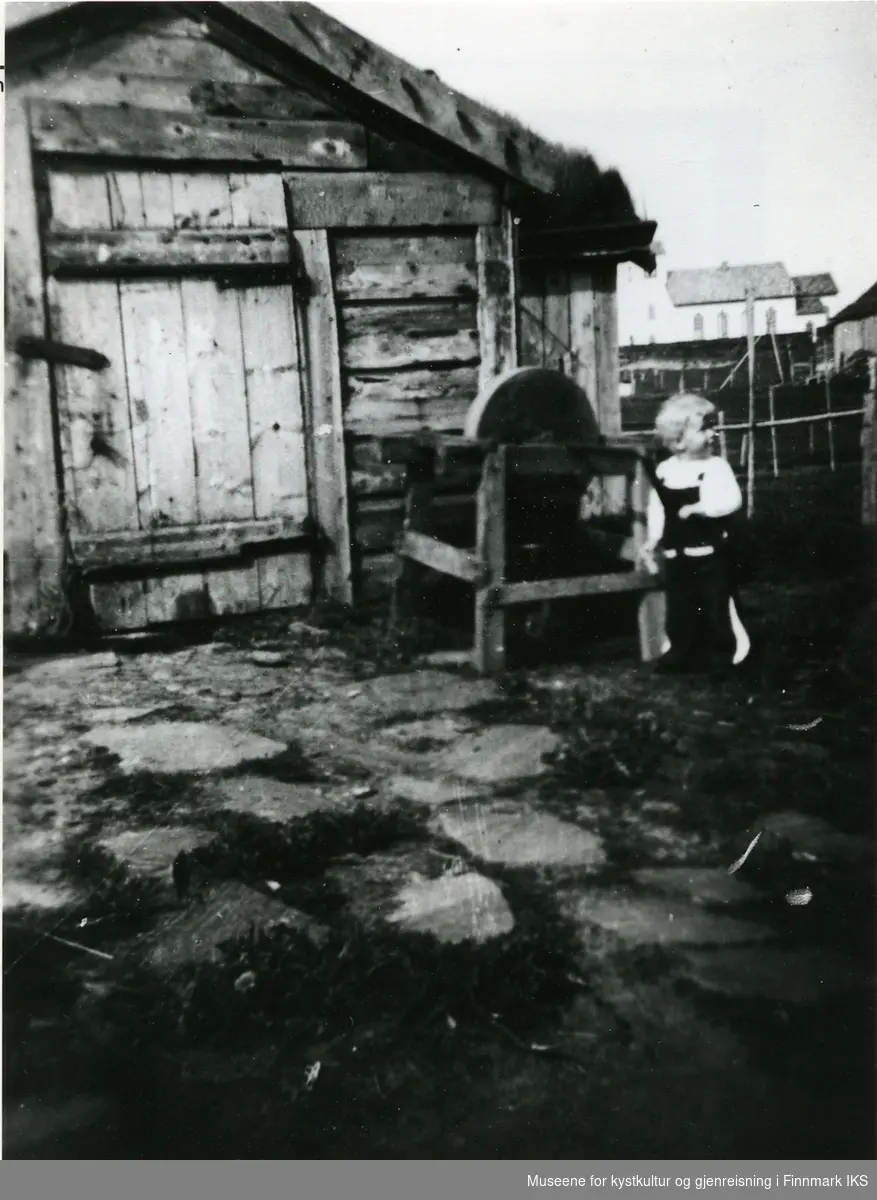 Et lite barn står ved en slipestein utenfor en liten bygning. Gamvik Kirke i bakgrunnen. Antatt 1920.