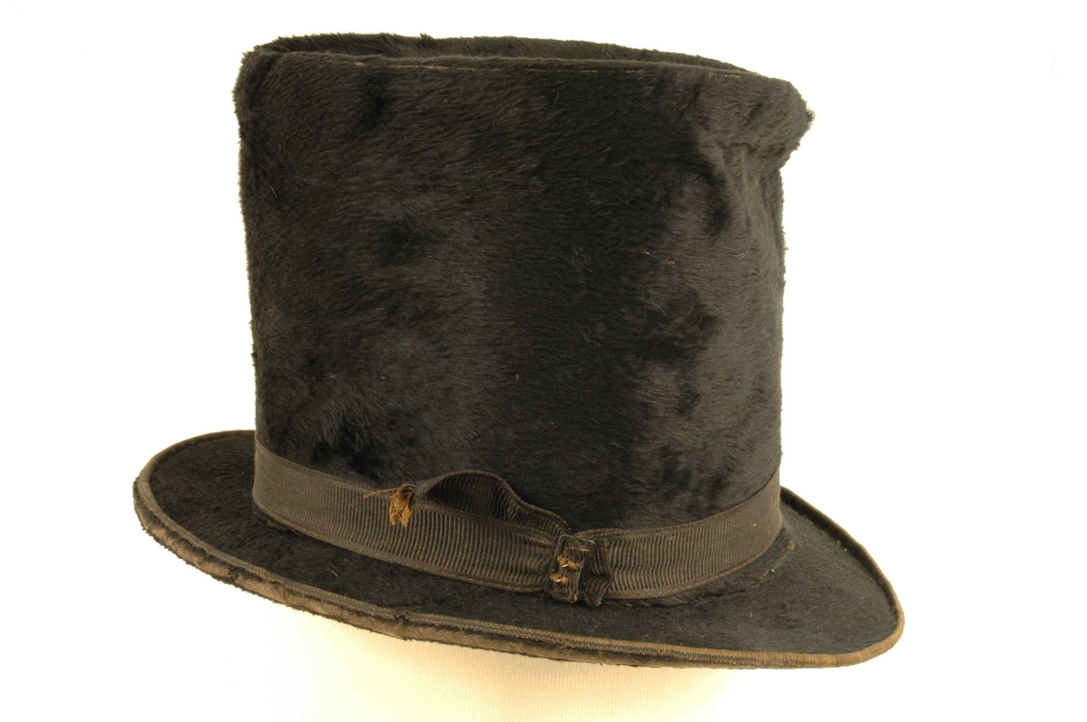 Svart flosshatt med høy pull og svart silke hattebånd. Smal brem.