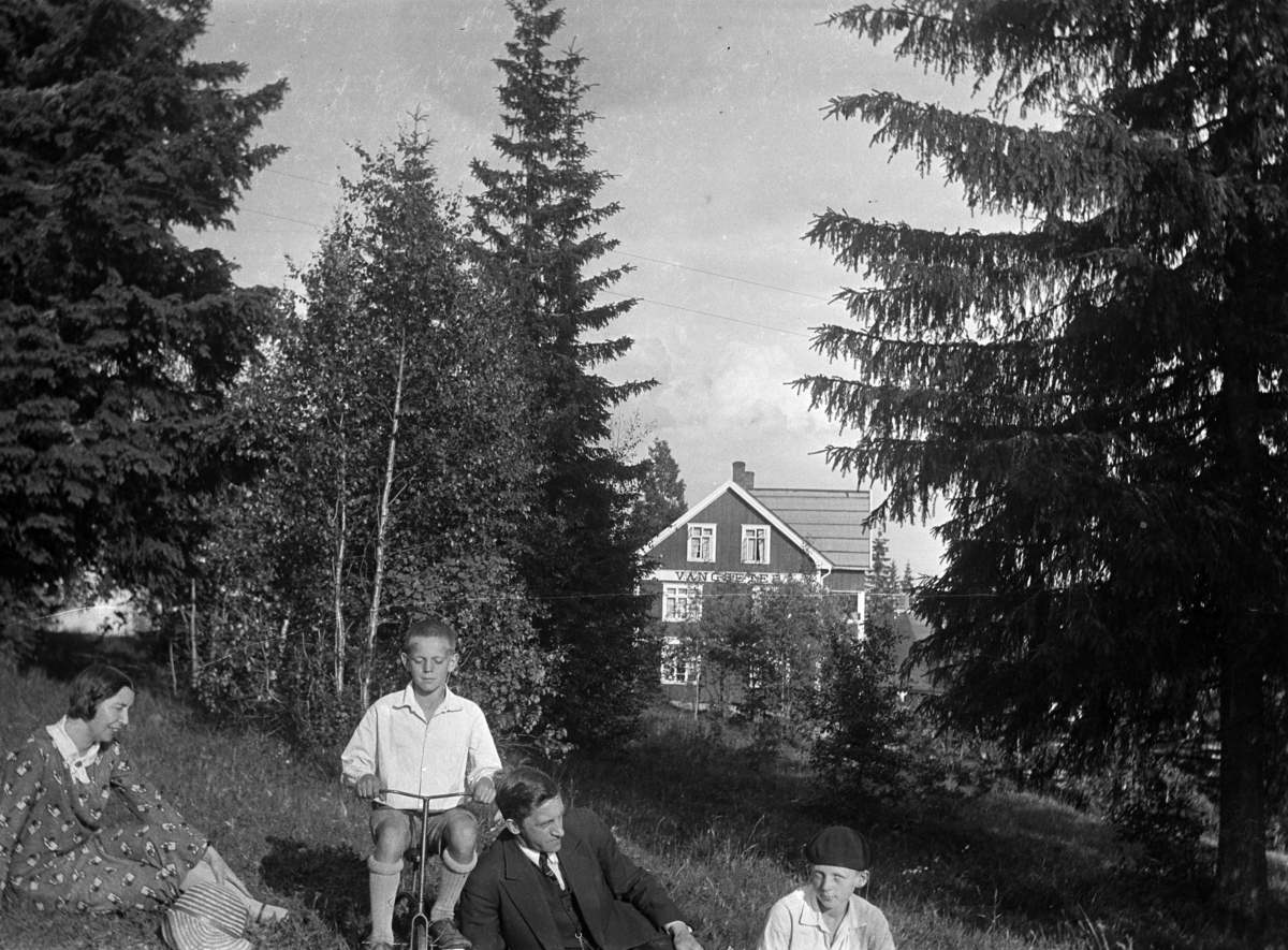 Ukjent kvinne, mann og to gutter i gresset, den ene på en trehjulssykkel. I bakgrunnen et hus med skilt Vangseter. 