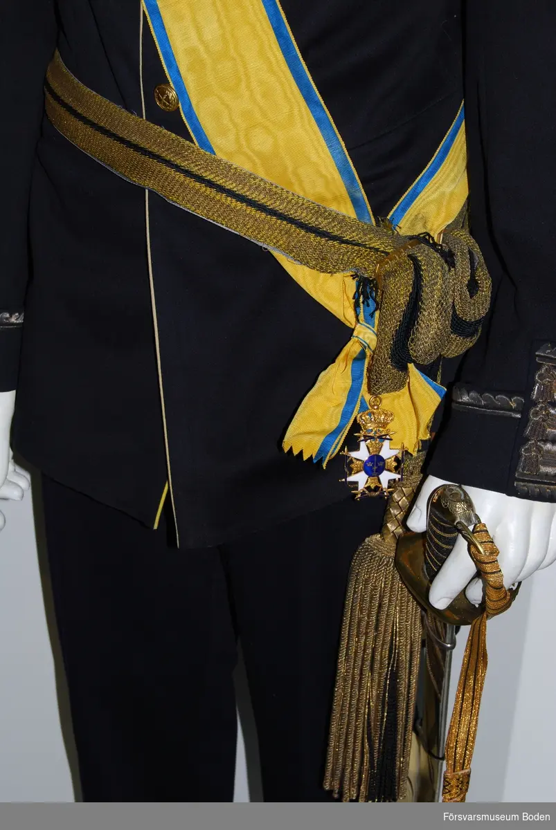 Virkat skärp i guldfärgat silke med blå rand i centrum. Två tofsar av blått silke med tjockare guldfrans  ytterst. Spänne av mässing för justering av längden.