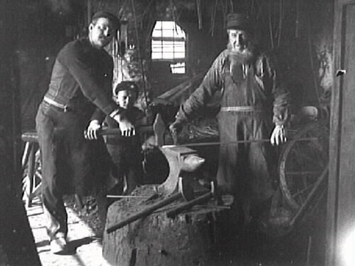 Interiör från en smedja. Två män, smeder, och en pojke vid står ett städ. Tre generationer i den gamla smedjan på Stomslätt eller "Smens" i Karl Gustav. Josef Andersson, med skägg till höger i bild. Mannen som håller släggan, i vänster bildkant, är Josefs son Emil. Pojken som står bakom Axel kan vara hans äldste son Axel. Bilden är tagen omkring 1912.