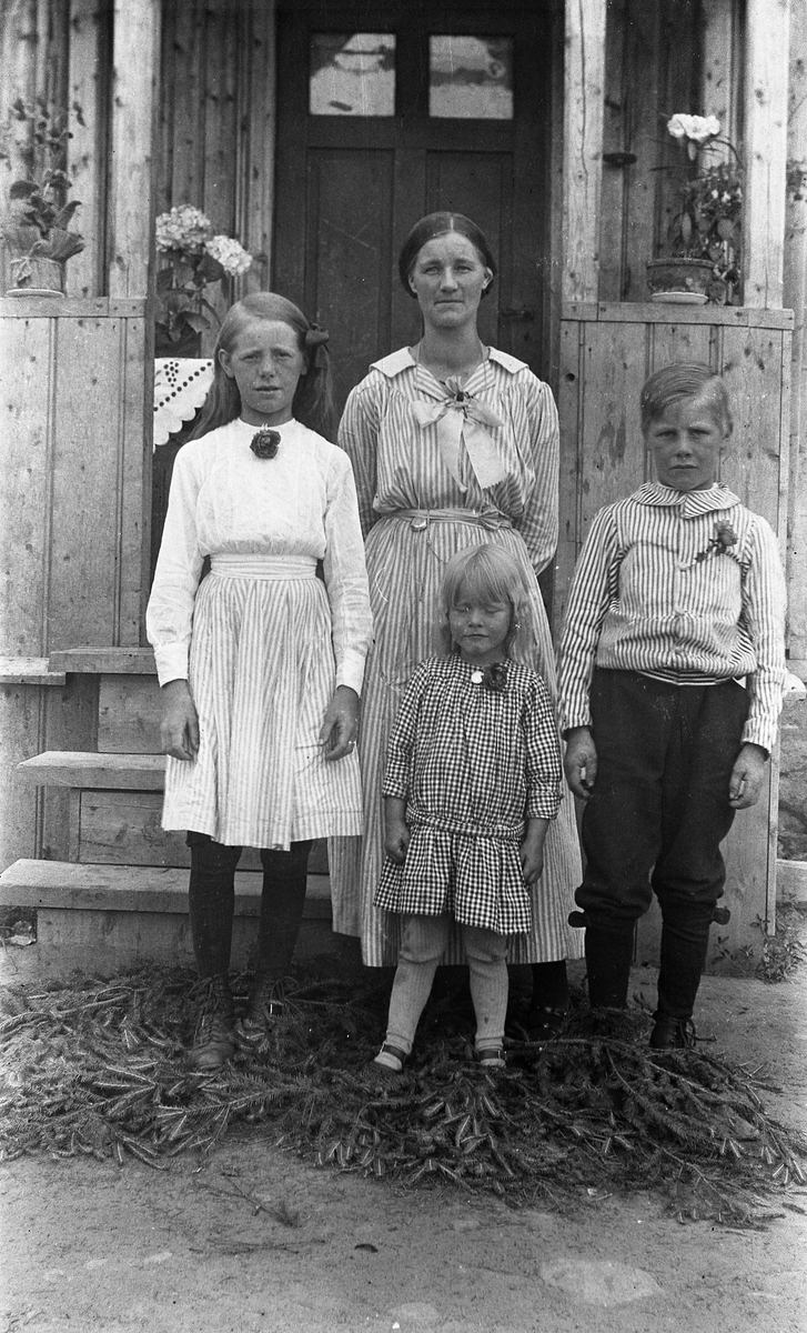 En voksen kvinne og tre barn foran inngangen til et våningshus. Bak fra venstre trolig Magnhild Tallerud, Olga Tallerud (gift Solberg), og Gunnar Tallerud. Disse tre var søsken.
Foran står trolig en yngre niese, Magnhild Tallerud (gift Nygård).