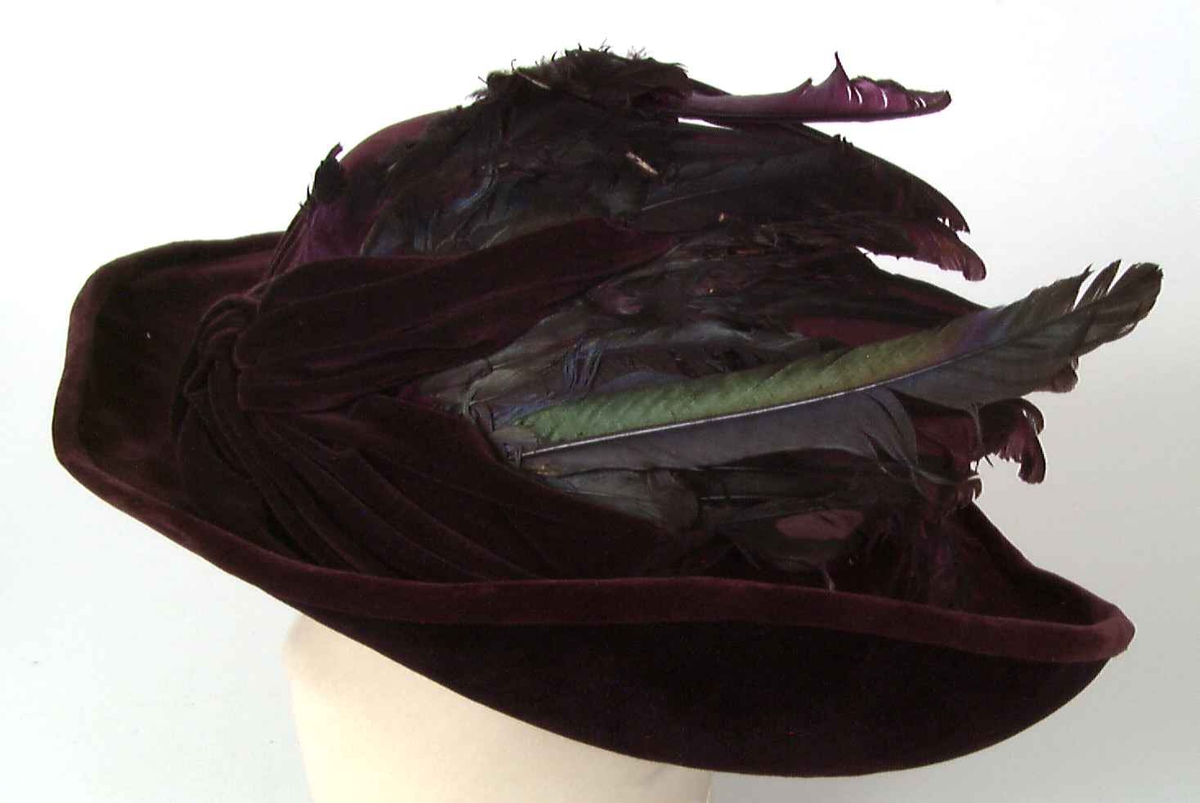 Vinrød hatt i fløyel med fiolett fløyel på brettets under side. Dekor av fjær.