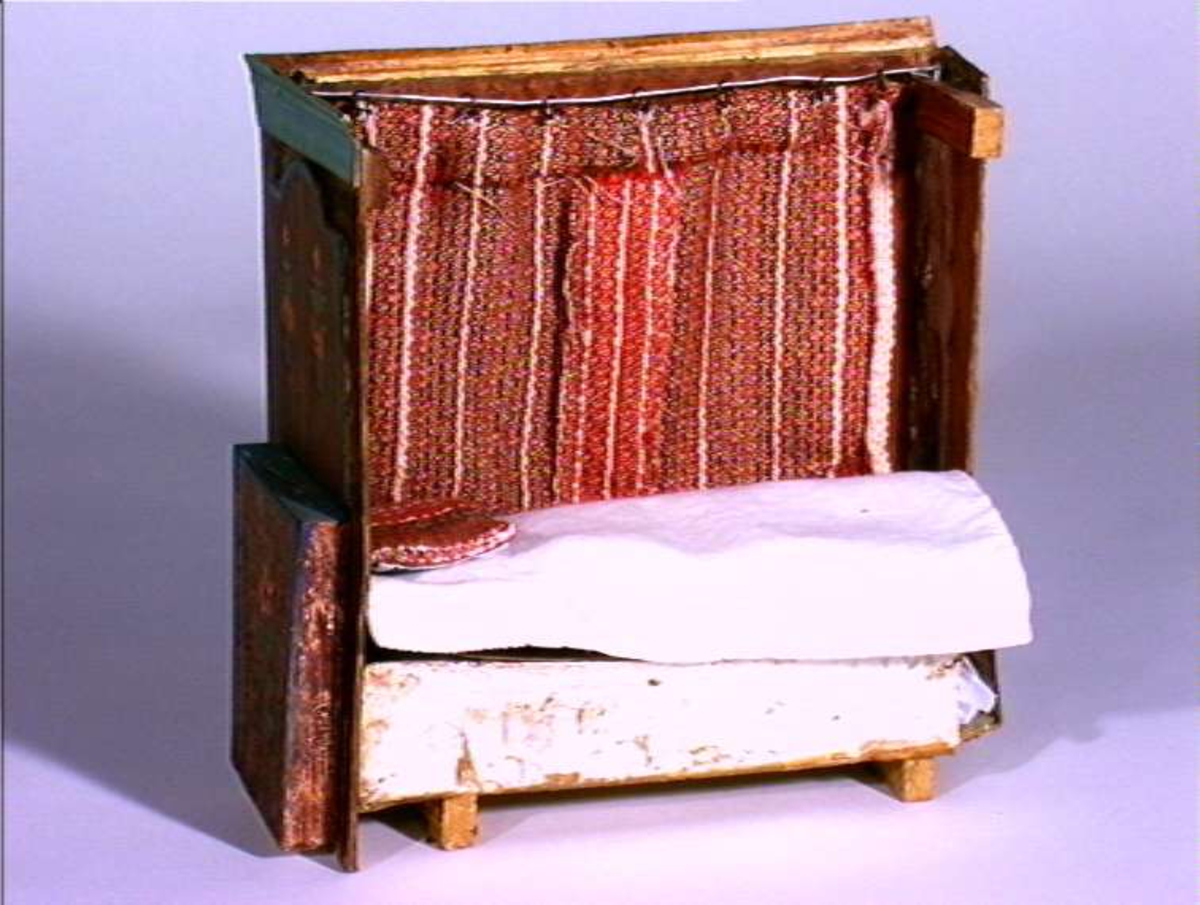 Dukkestue i form av svensk hytte med påmalte tømmerstokker, grunnmur og takstein i tegel. En vegg kan løftes bort og taket kan også åpnes. Stuen har tre stk vinduer med påmalte blysprosser. Inne i stuen er peis, bord og en dukke, en man i svensk folkedrakt med fele, fastmontert. På veggen henger et kurbitsdekorert maleri av Carl Johan.