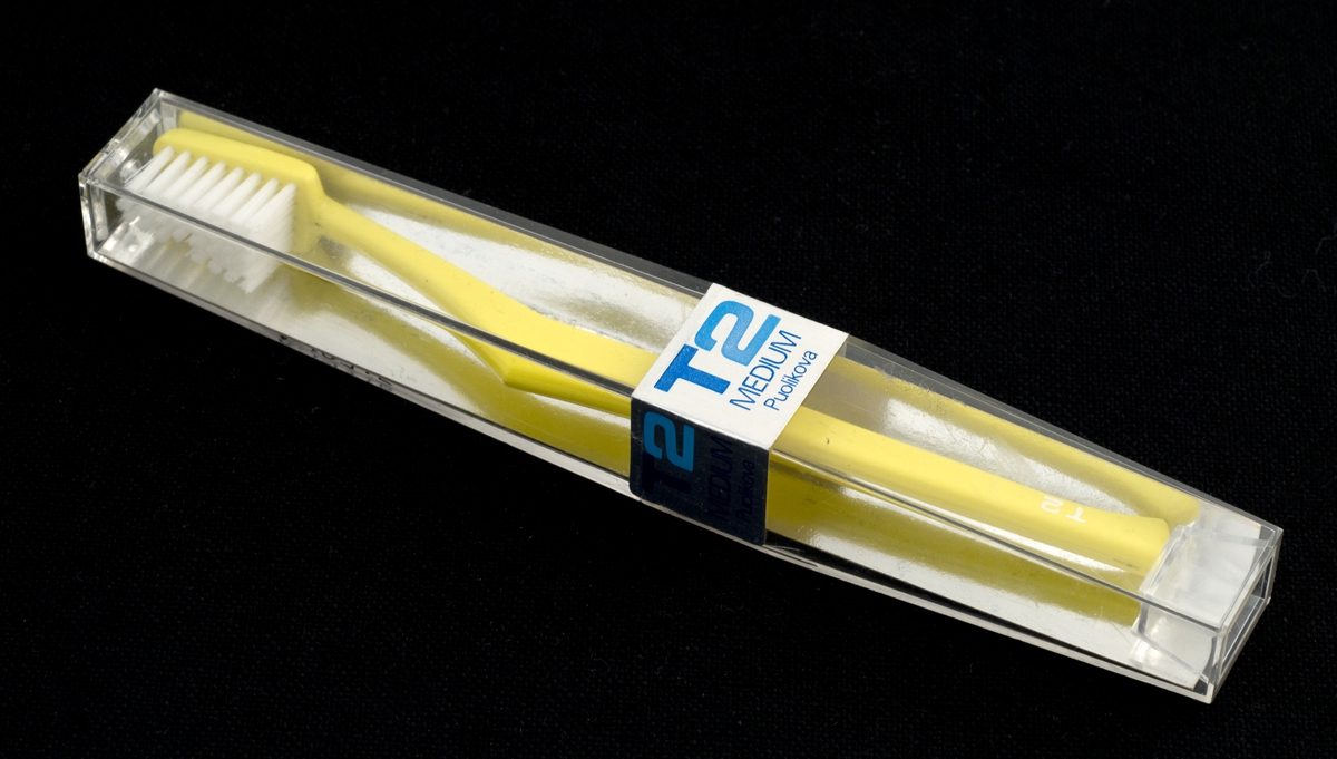 Tannbørste i uåpnet orginalpakning. Emballasje av gjennomsiktig plast. Inneholder tegning av en tannbørste i blått og firkanter i hvitt, mørkeblått og lyseblått.