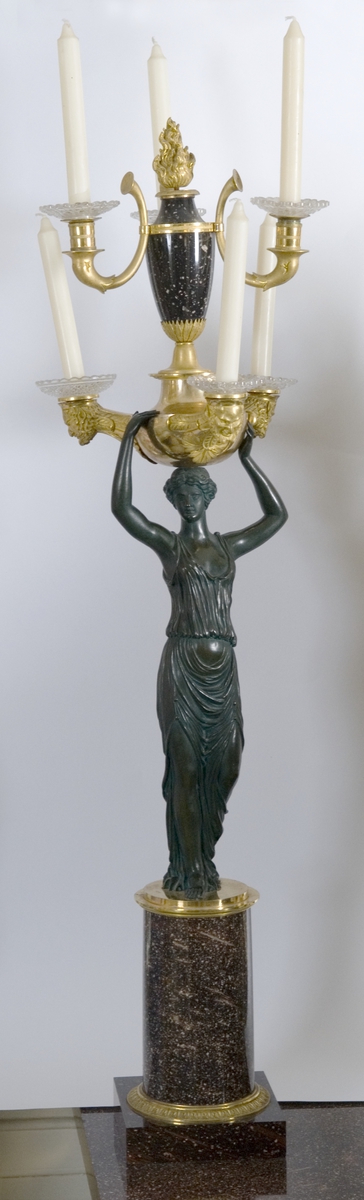 Kandelaber, porfyr, bronse forgylt og bronse mørk. Fotplate firkantet, sokkel rund, på den står kvinnefigur i klasisk gevant, hun bærer romersk lampe med tre lysholdere, over den urne med tre lysholdere.