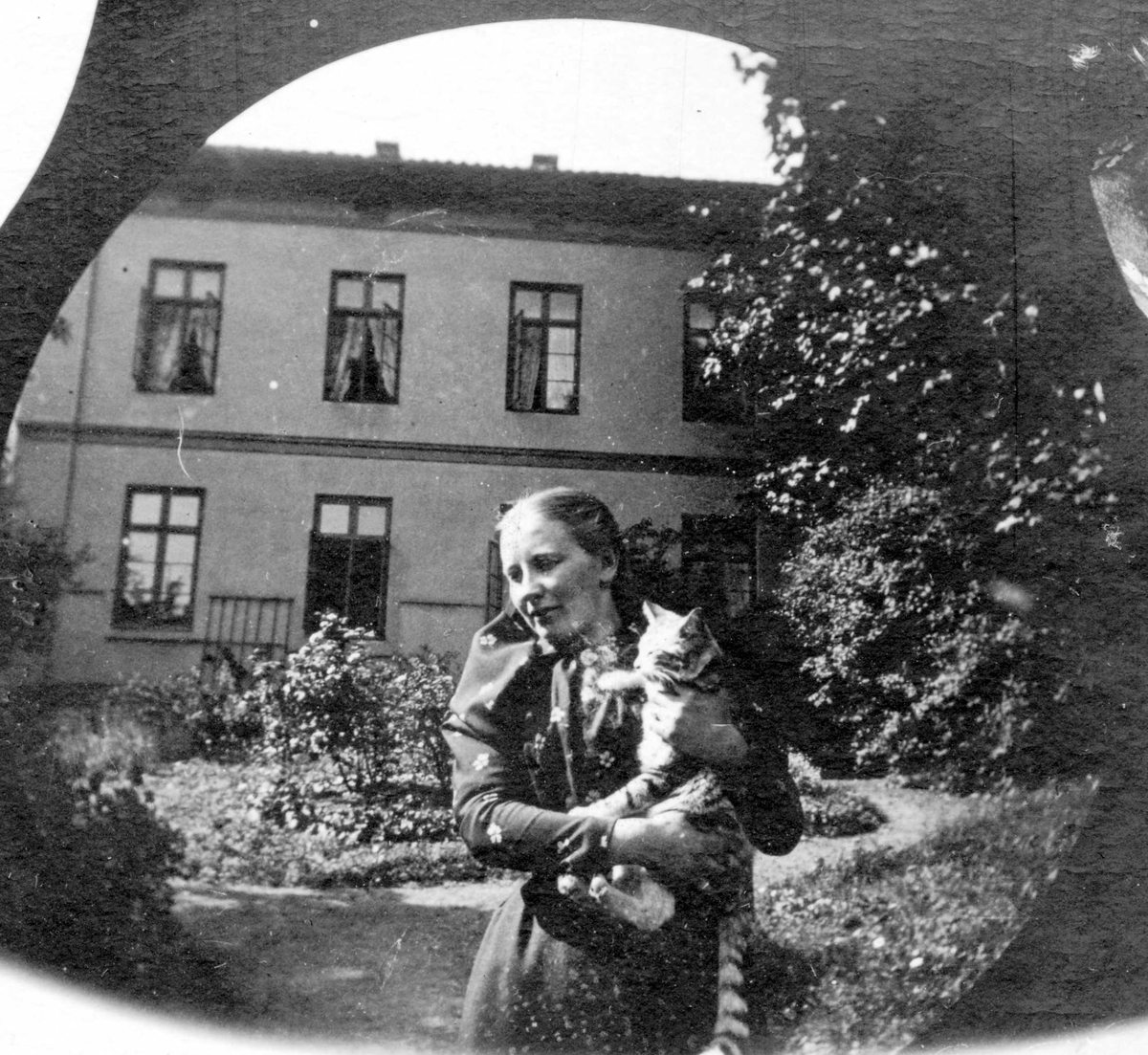 Professor Blytts stuepike og katt. Kvinne med stipet katt på armen i hage forann toetasjers hus, Oslo.