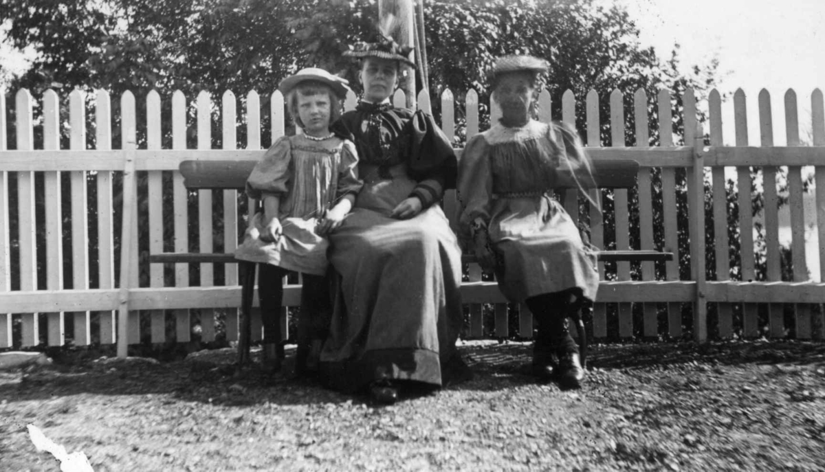 Div. drakt, Norge 1908-10. En liten pike sittende på en benk med to kvinner. Begge i lange kjoler/skjørt  og med hatt. Piken i kjole og med hatt.