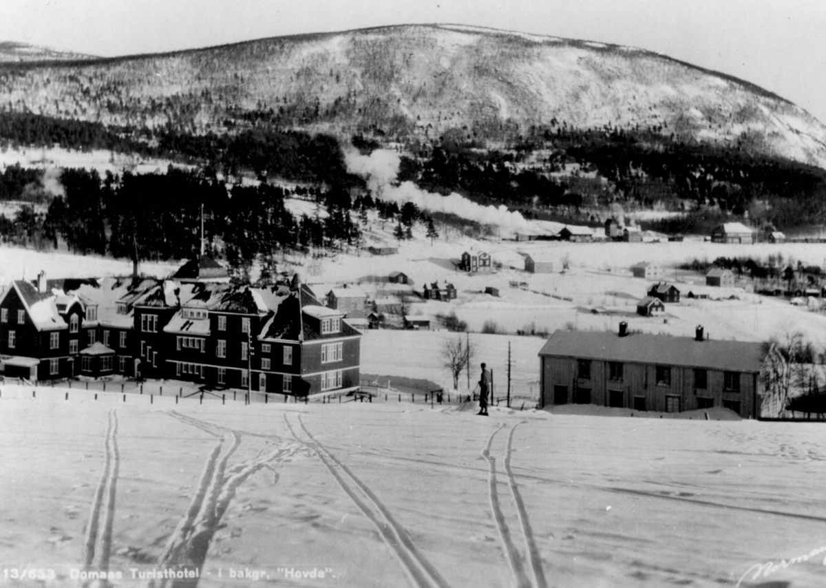 Avfotografert postkort. Skispor og en mannlig skiløper foran Domaas turisthotell. I bakgrunnen sees bebyggelse og fjellet Hovda.