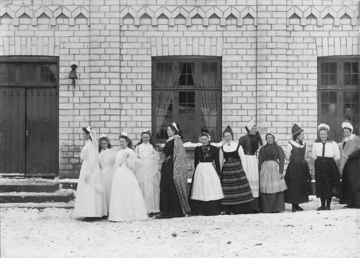 Kvinner i drakter, to av dem i bryllupsskjoler står i rekke foran en bygning. Muligens svensk.
