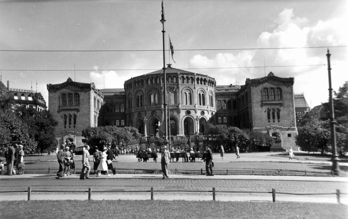Stortinget og Eidsvolls Plass, Oslo 1939. Fotografert fra Rosenkrantz' gate. Flagg på Stortingsbygningen. Menn og kvinner sittende på benker og spaserende foran Stortinget.  sSpaserende foran.
