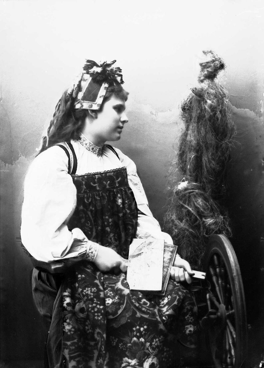 Studioportrett av kvinne i drakt og skaut, som sitter ved en rokk og karder. Bakgrunnen er malt.