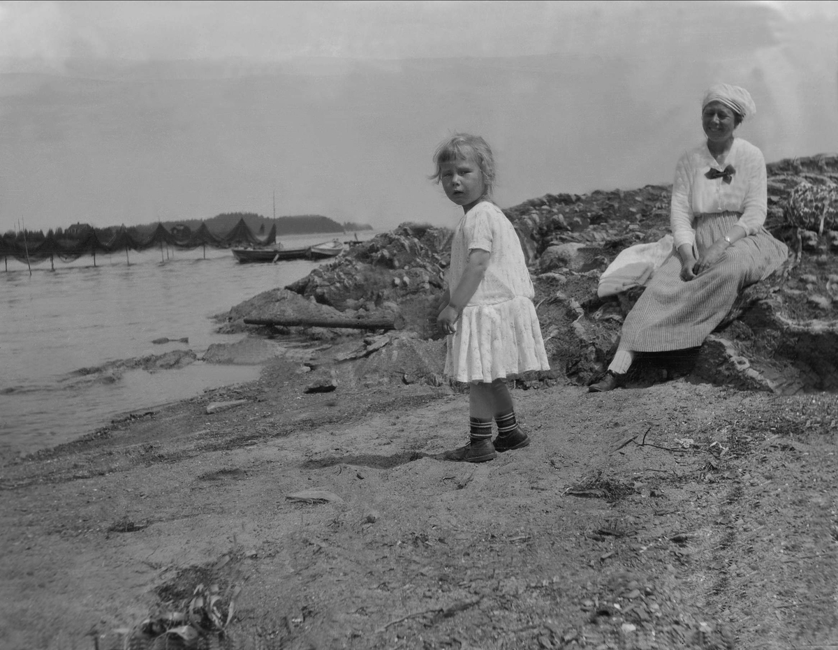 En kvinne sitter på svaberget og ser på en jente som står på en strand. Robsahm og Lund.