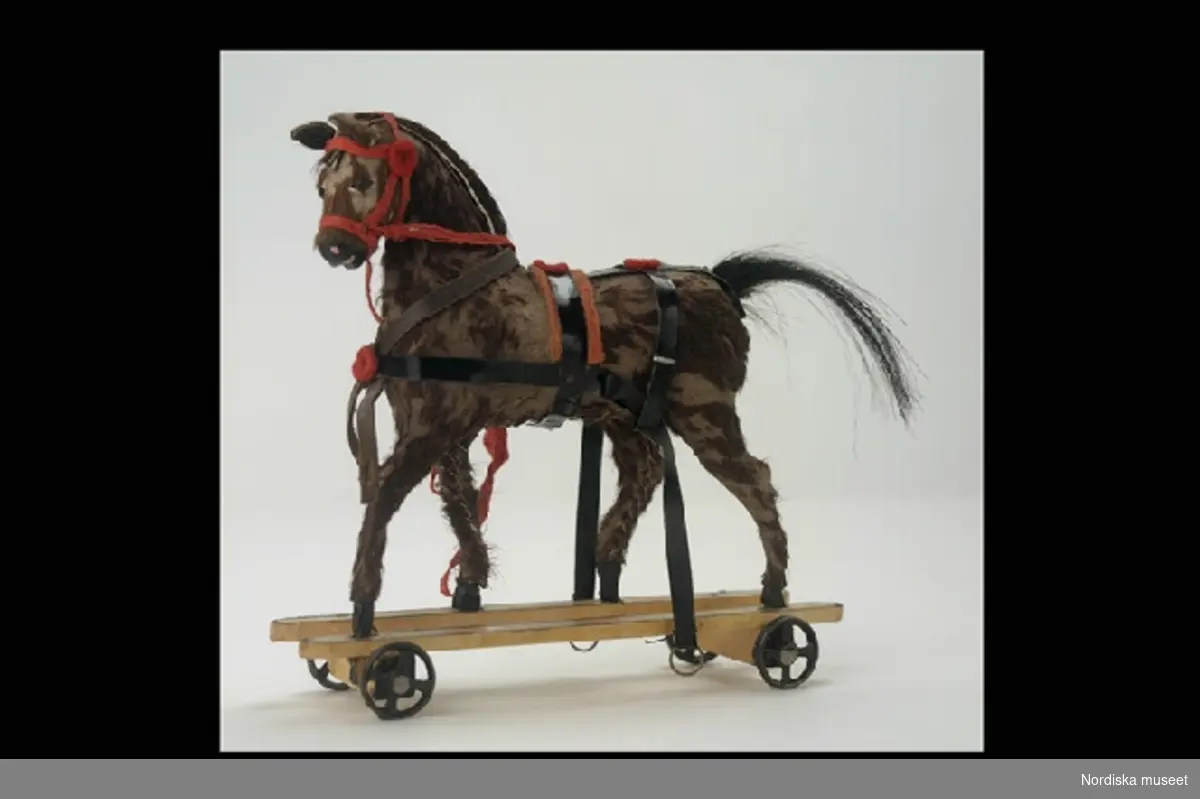 Inventering Sesam 1996-1999:
Häst L 15,5 cm, H 15,5 cm
Trilla L  35  cm  B 11  cm
Häst och trilla.
Häst av skinn med brun päls på hård stomme. Ögon av metallnubb. Svans av svart tagel. Seldon av svart vaxduk och röd filt, träns och töm av röda band. Hästen fäst på underrede av trä med fyra små svarta metallhjul.
Trilla av trä, gulmålad med röda ränder, två säten, hjul med ekrar, främre hjulparet svängbart, två skaklar.
Givarinnan dotter till leksakshandlare W A Bandel, Västerlånggatan, Stockholm. 
Birgitta Martinius mars 1998