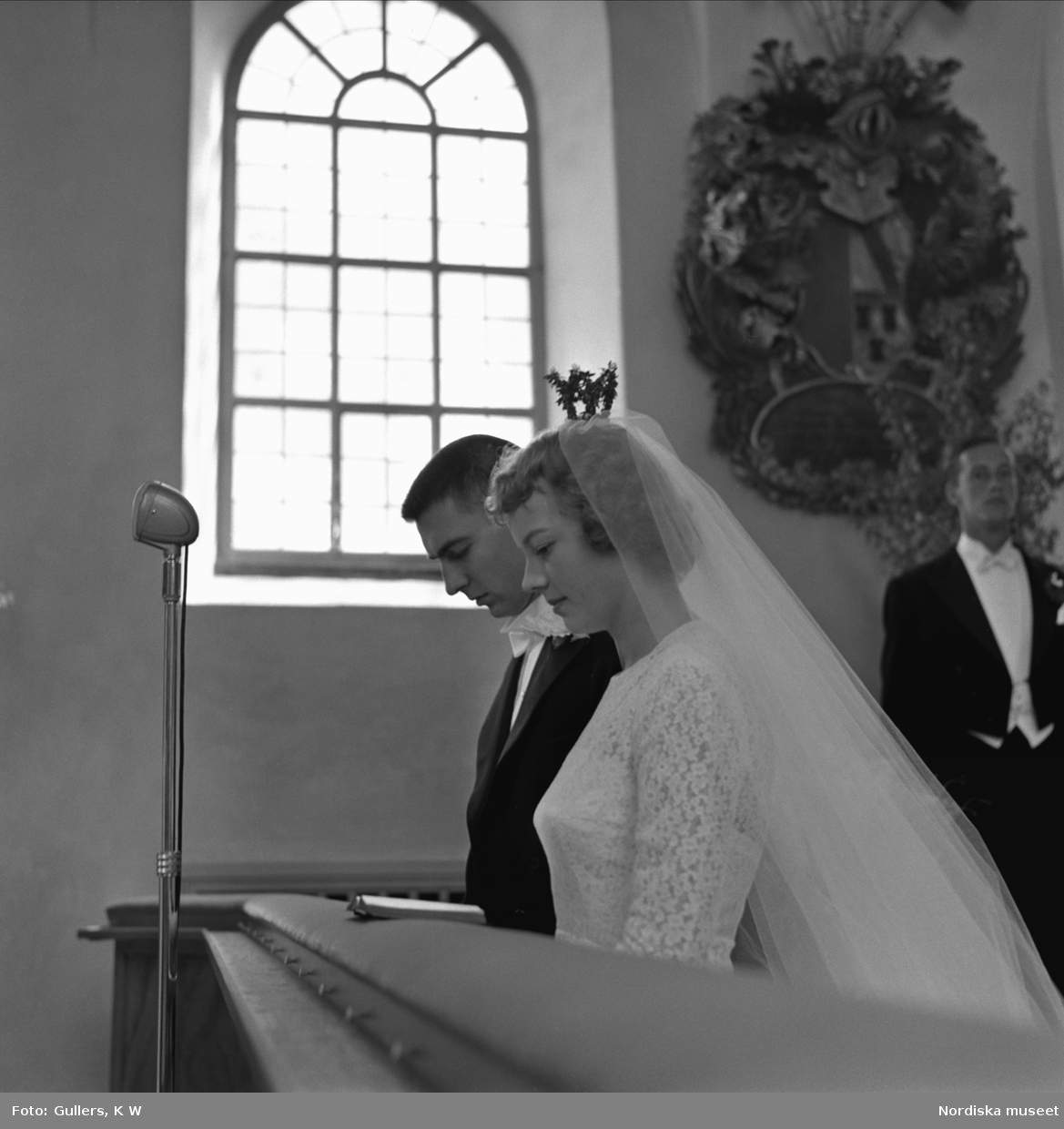 Bröllop, bild av ett bröllopspar som knäböjer i en kyrka. Bruden iklädd vit klänning, brudslöja och liten krona. Brudgummen iklädd frack.