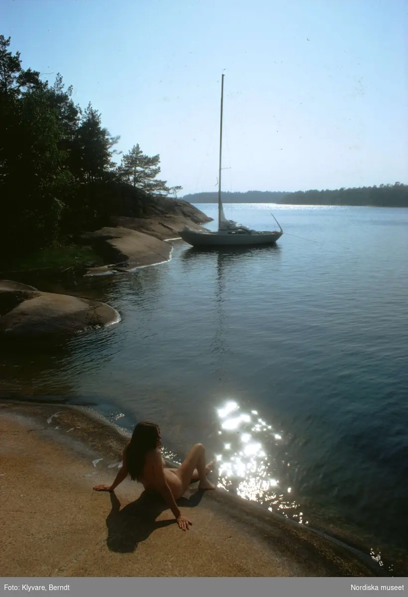 Naken kvinna på klippa i Stockholms skärgård. I fonden en segelbåt