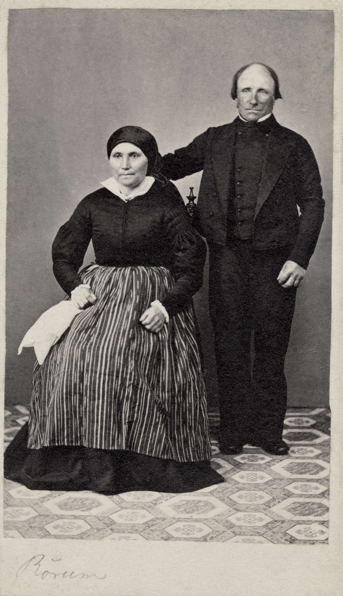 Parporträtt, kvinnan iklädd sjalett och randigt förkläde sitter, mannen står vid hennes sida. Skåne, Albo härad, Rörums socken. Nordiska museet inv.nr. 18533B.
