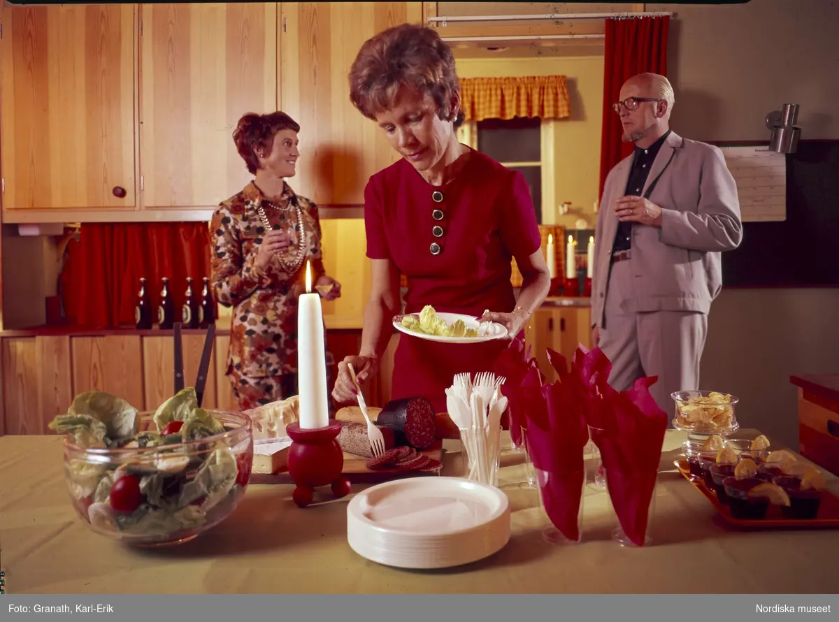 Traditioner. Jul. Köksinteriör; en kvinna plockar mat på en tallrik. En man och en kvinna pratar i bakgrunden.