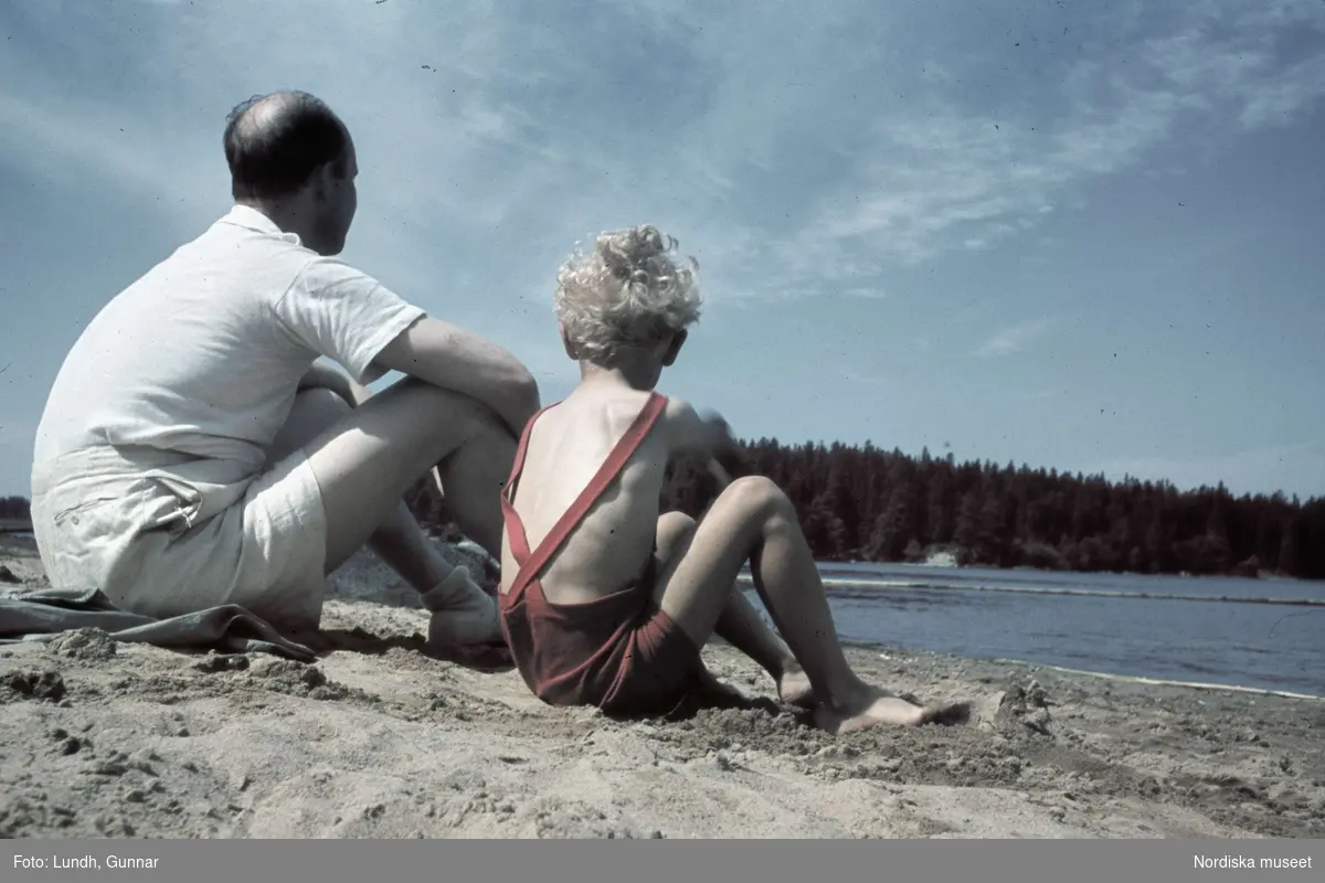 En man och en pojke sitter på sandstrand. Mälarhöjden, Stockholm