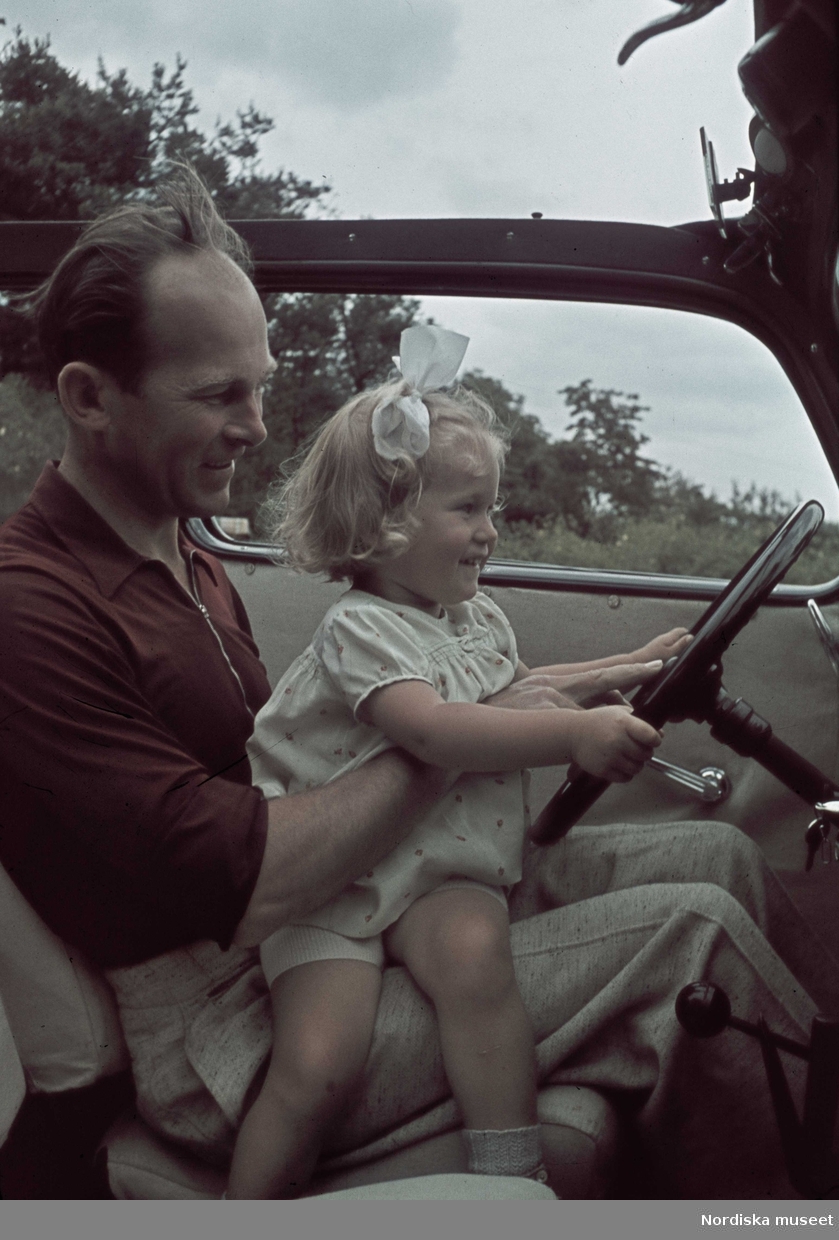 En man sitter i en bil med en liten flicka i knät som håller i ratten.
