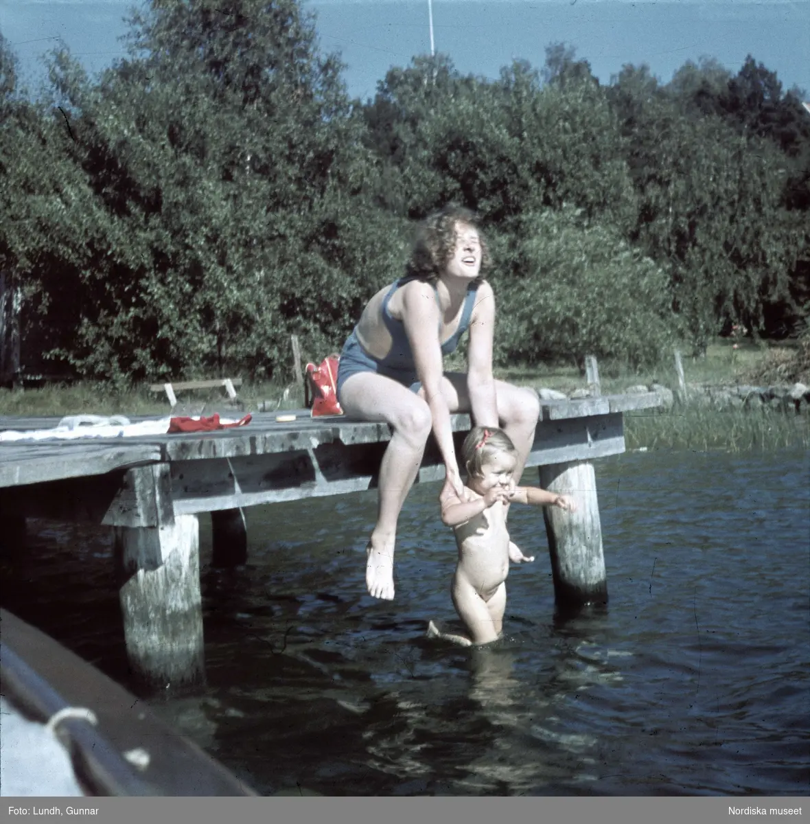 Kvinna doppar ner en flicka i vattnet från en brygga.