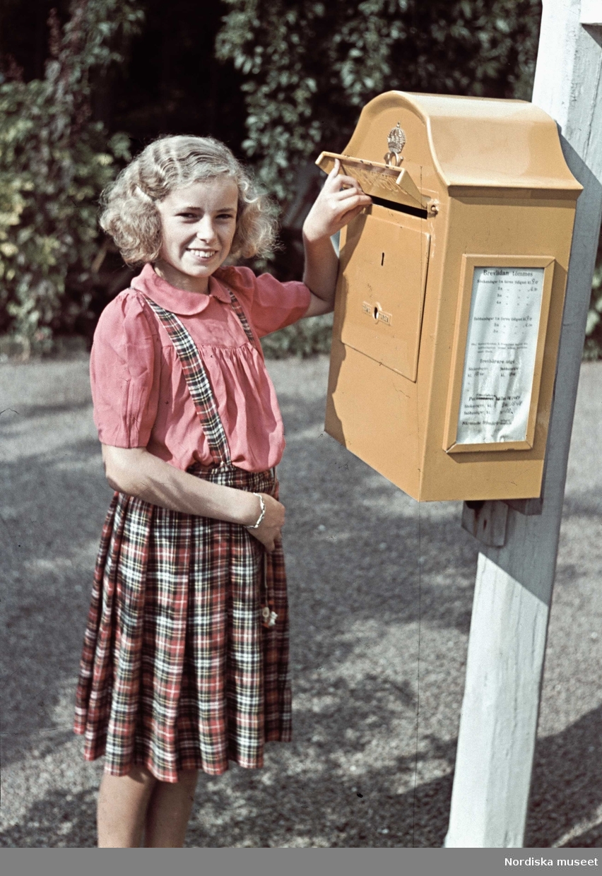Flicka i röd blus och rutig hängselkjol vid en brevlåda. Troligen fotograf Gunnar Lundhs dotter Jytte.
