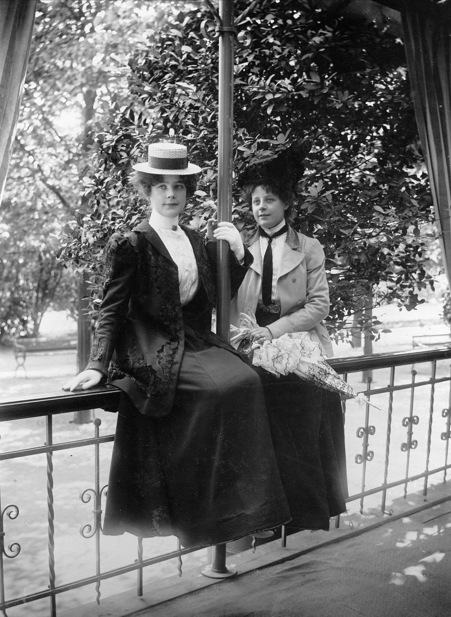 Två unga kvinnor i dräkt och hatt sittande på räcke.