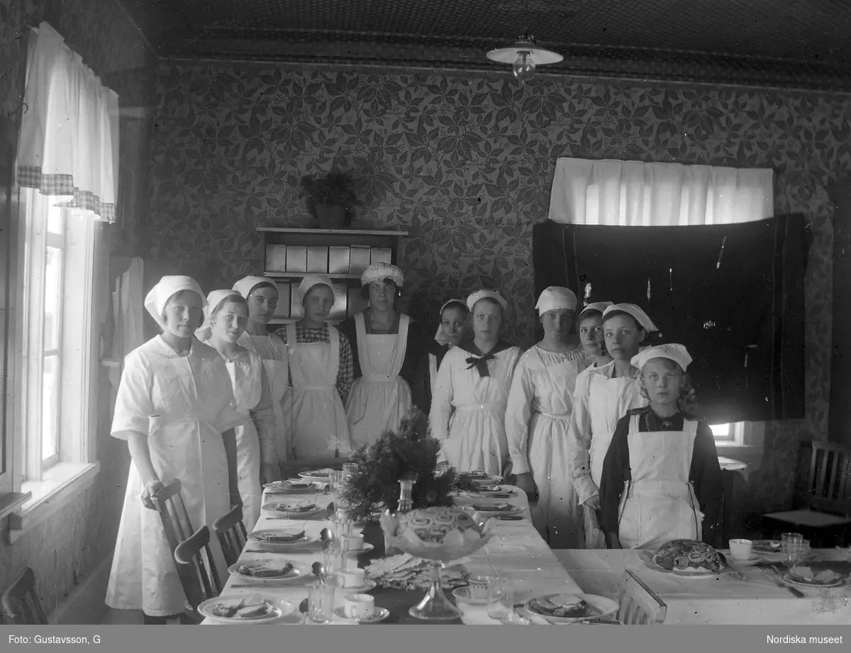 Grupppbild. Elva kvinnor i vita förkläden och mössor bakom ett dukat bord. Stormönstrade tapeter, även taket har tapeter.