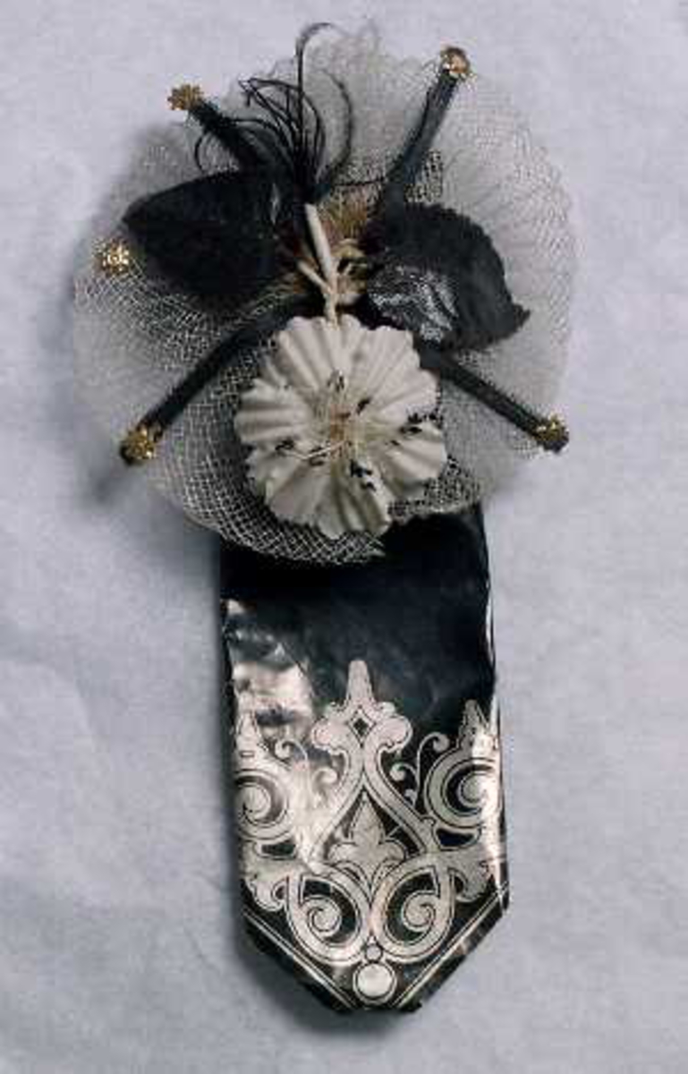 Begravningskaramell av svart glanspapper med silvermönster. Prydd med flor av vit tyll och blommor av vitt tyg med blad av svart tyg.

