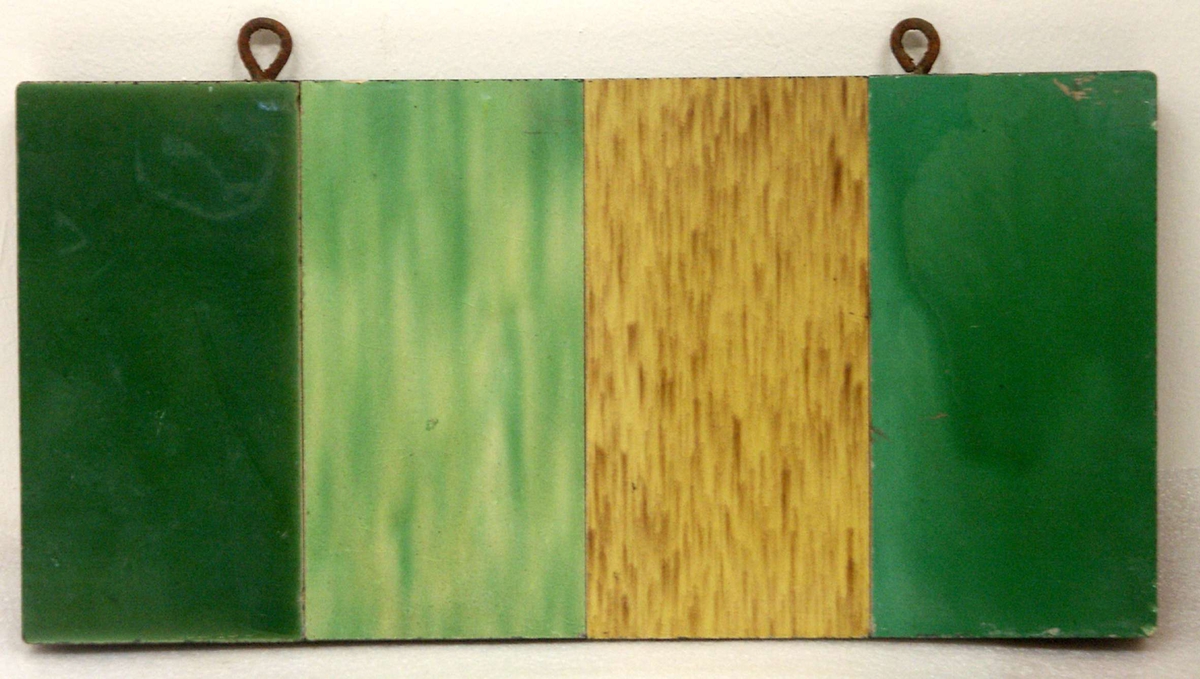 Platta av gips, på vilken fyra kakelprover i en gul och tre gröna nyanser är monterade. Svartmålad på kanten och större delen av baksidan. Upptill två öglor av järn.