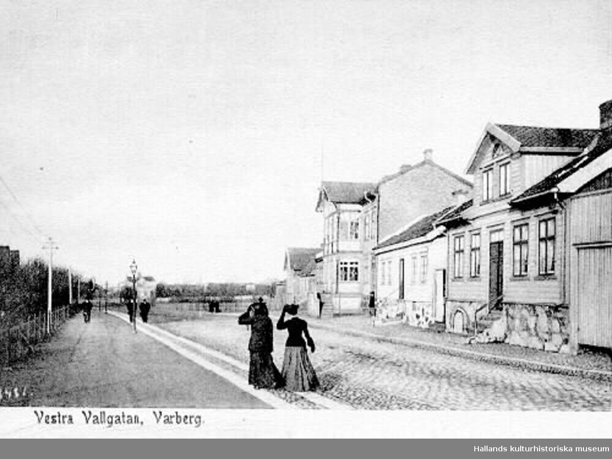 Brevkort, "Vestra Vallgatan, Varberg" (meddelandet skrivs på den tomma ytan på framsidan). Bilden är tagen mot norr så till höger syns kvarteret Berget och längs vänster sida ligger Järnvägsparken. Två damer promenerar och håller i hattarna. De vita stolparna till höger är Hugo Gerlachs elstolpar (uppfördes 1892) som går från hans Mekaniska verkstad, via Järnvägs- och Engelska parkerna till hans bostad i Franckalyckan. Stadens gatlyktor står på gångstråket.