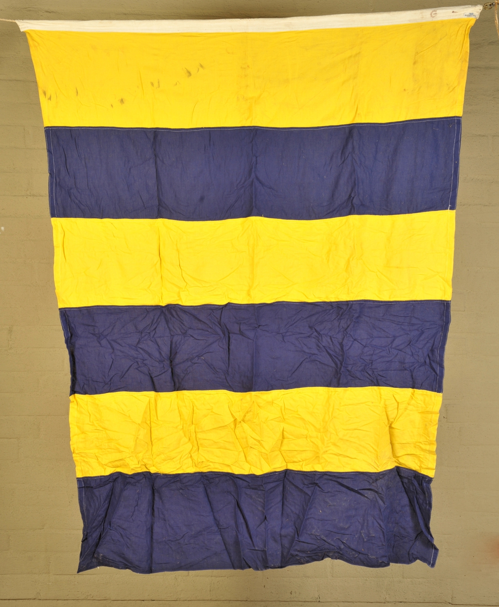 Signalflagg med 6 vertikale striper: gul, blå, gul, blå, gul, blå. Viser bokstaven G.