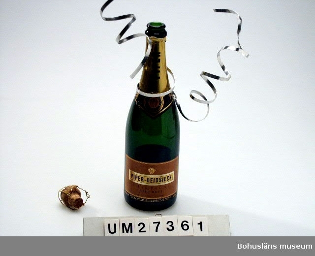 Champagneflaska (:1) i grönt glas med pappersetikett om hals och buk, 750 ml. Serpentin av silverfärgad mjuk metall virad runt flaskhalsen.
Kork (:2) med text "Piper" och tillslutning av virad metalltråd.
Flaskan är brukad i samband med nyårsaftonsupé för fyra personer.

Se även Bilagepärmen UM27361 för exempel på varningsartikel som publicerades i pressen i januari 2000 med anledning av exploderande cider- och champagneflaskor.

För information om Millennieinsamlingen, se UM27360.