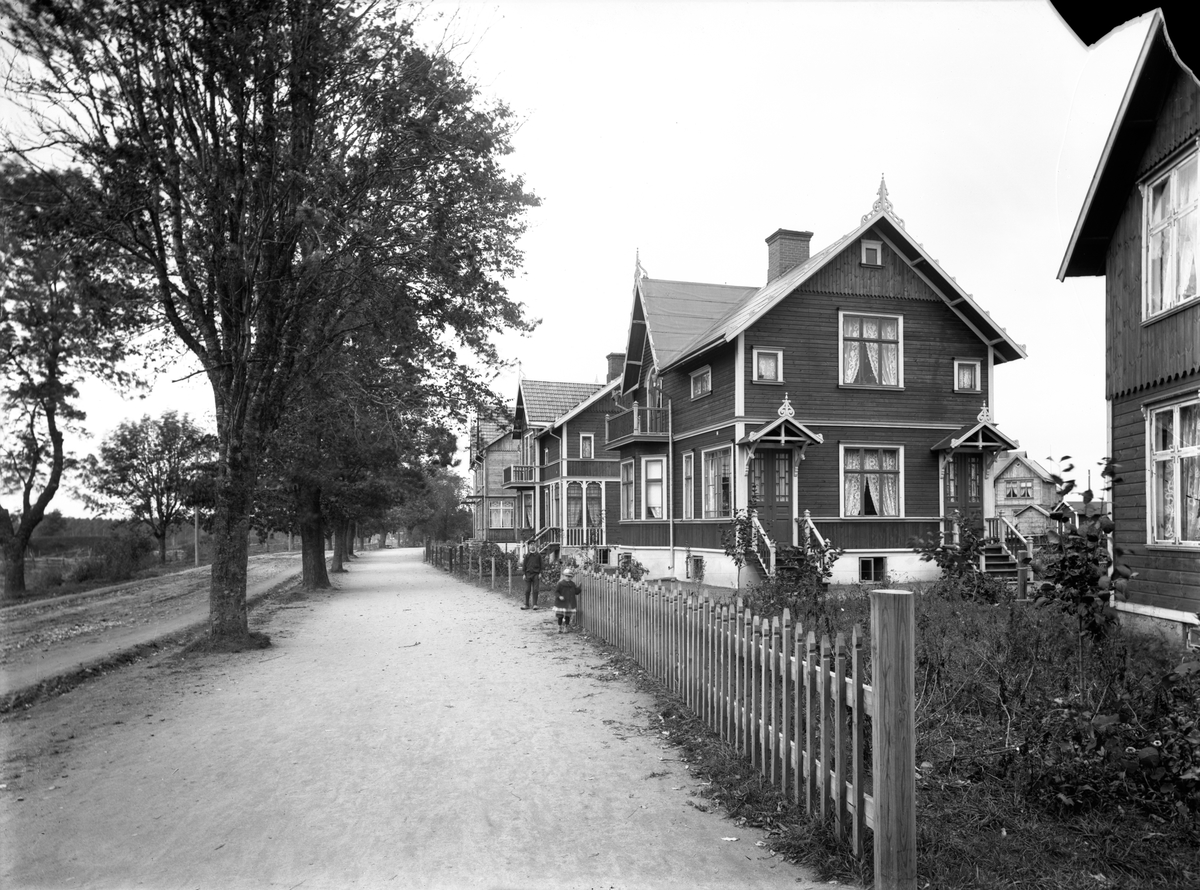 Jönköpingsvägen i Huskvarna år 1908. Tvåvåningsvillor i trä ligger utmed den trädkantade vägen. Två barn står vid staketet på trottoaren.