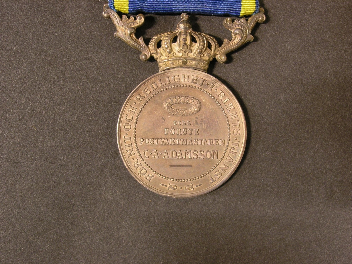 Medalj tilldelad förste postvaktmästaren C A Adamsson för
"Nit och redlighet i rikets tjänst". Medaljen är av silver med ett
blått och gult sidenband. Medaljen skall bäras på bröstet. Medaljen
visar på åtsidan kung Gustaf V i profil åt vänster. Frånsidan har en
lagerkrans över inskriptionen "TILL FÖRSTE POSTVAKTMÄSTAREN C A
ADAMSSON" i fyra rader. Medaljens ovansida har en kunglig krona och
två lagerkvistar som bildar en upphängningsanordning för sidenbandet.