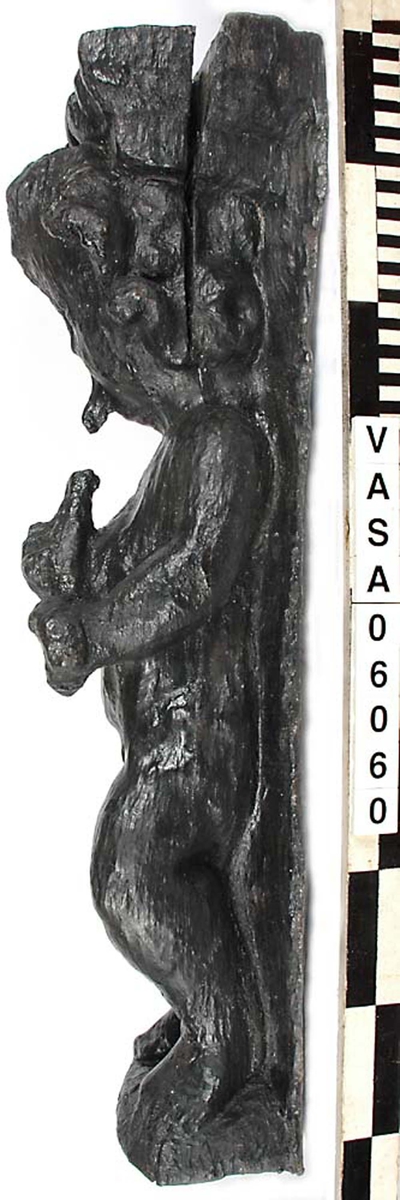 Skulptur föreställande en barnfigur som spelar på en sinka (ett äldre blåsinstrument med grepphål som blockflöjten men ett munstycke som liknar trumpetens).
Huvudet är vridet något åt höger. Kroppstyngden vilar på höger ben medan vänster ben är böjt. Instrumentet hålls med båda händerna. Skulpturen förefaller delvis draperad i en mantel eller dylikt som skymtar vid hans högra sida. Ovanför hjässan syns en något snedställd äggstavslist. Figuren står på ett minimalt underlag som troligen avser att föreställa ett stycke naturlig mark. Skulpturens baksidan är slät.
Skulpturen är kraftigt sliten.

Text in English: A sculpture of a child playing a sinka.
The weight of the body rests on the right leg while the left is bent and the head is turned slightly to the right.. The instrument is held in both hands. The figure seems to be partly draped in a cloak or the like, which conceals his right side. Just above the head the back board comes forward at right angles and the front of the board is decorated with an egg  mould design. The sculpture stands on a small flat plinth. The back of the sculpture is smooth.
The sculpture is very worn.