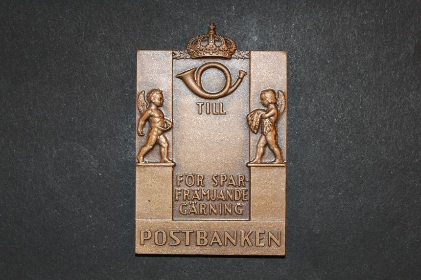 Plakett, tillverkad i brons, utformad av konstnär KarlHultström. Rektangulär. Plaketten visar överst ett krönt postemblemav 1937 års modell. Under emblemet står text "TILL .......FÖR SPARFRÄMJANDE GÄRNING". På bägge sidor om textfältet står två bevingade människor föreställande sådden och skörden. Underst stårtext "SVERIGES POSTSPARBANK" flankerad av två uppsättningar avTre Kronor. Plaketten är märkt med tillverkarens namn (Sporrong).
