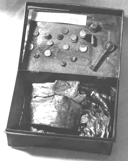 Kassaskrin av bleckplåt, innehållande sedlar och mynt i förbrännt skick. Med nyckel (PM 2858) och mynt (PM 2859).