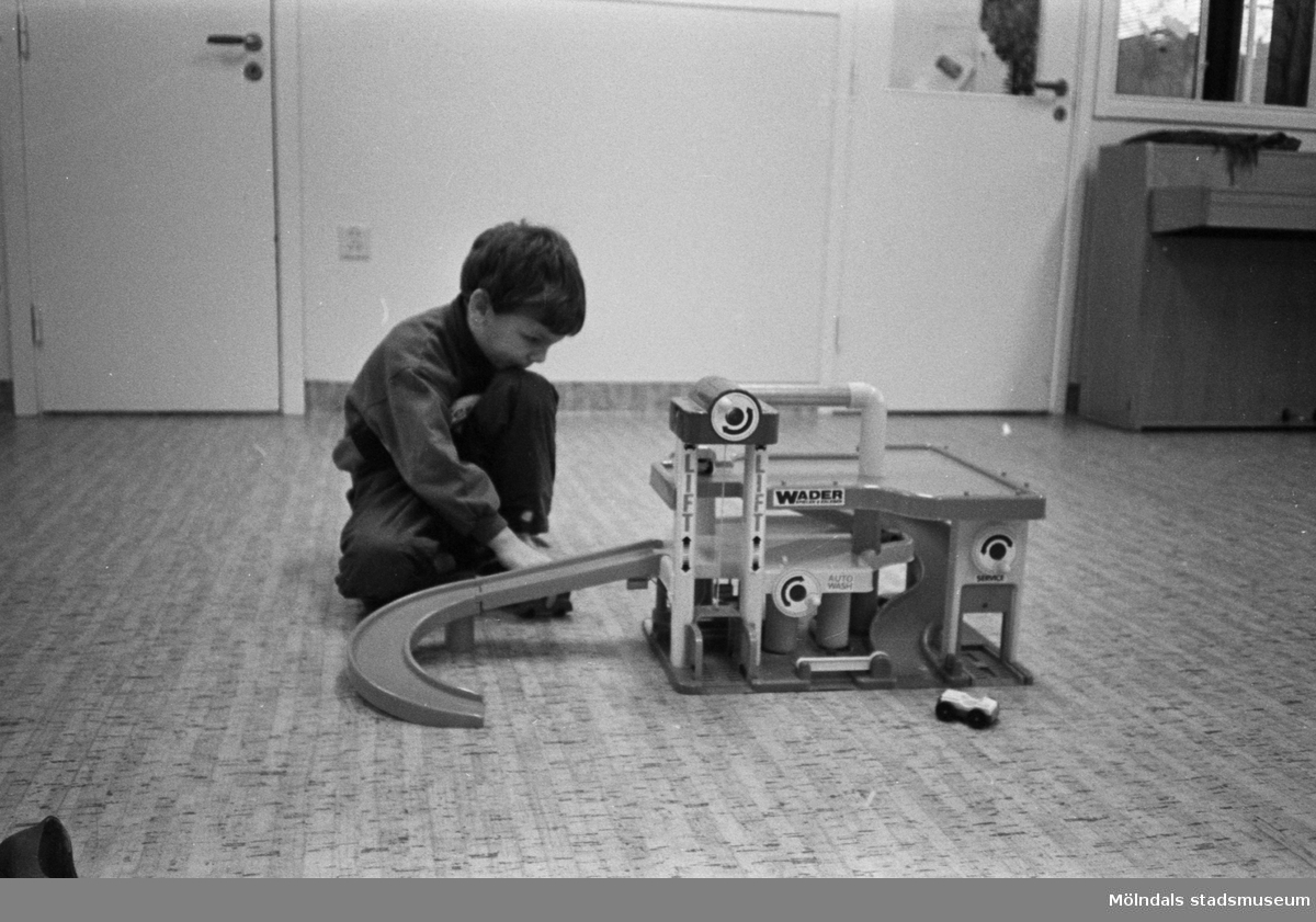 En pojke sitter och leker med ett Wader-garage utrustat med bensinpumpar, hiss, tvätt- och servicestation samt en lång avfart för tillhörande leksaksbilar. Han sitter på ett korkliknande linolium-golv. I bakgrunden till vänster syns en stängt dörr. Till höger står ett piano.