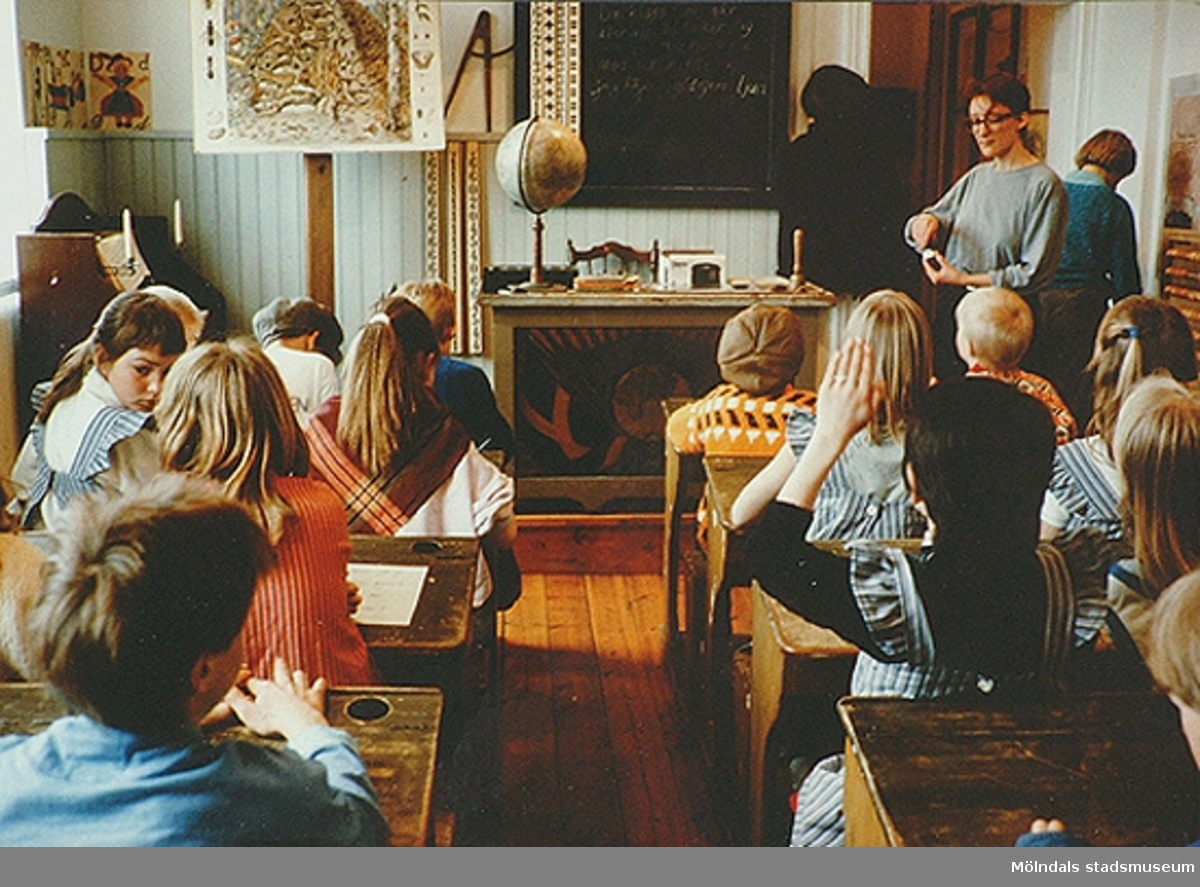Barn som sitter i skolbänkar och skolsal av äldre modell. Mölndals museum, 1990-tal.